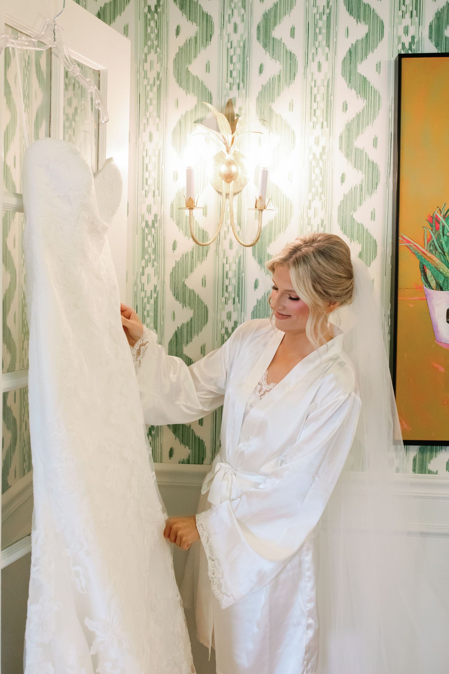 Bride in white satin robe admiring her wedding gown hanging on a door in an elegant green wallpapered room.