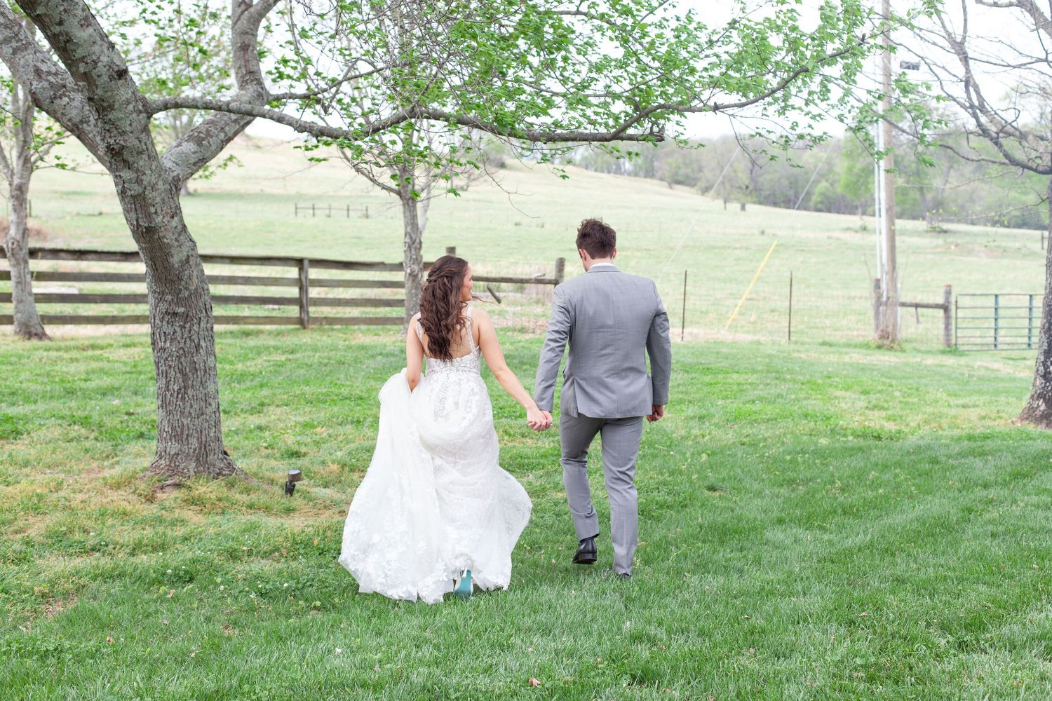 Bride and groom walking hand in hand across lush green grass near a wooden fence at an outdoor wedding venue.