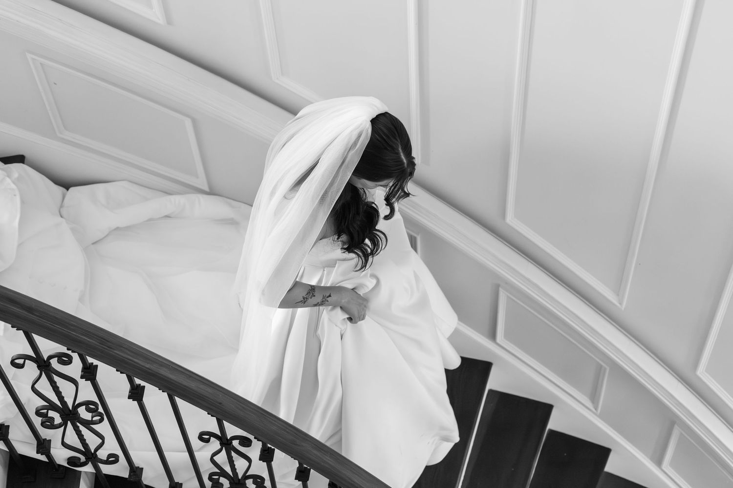 Black and white photo of a bride in a flowing gown and veil descending an elegant curved staircase.