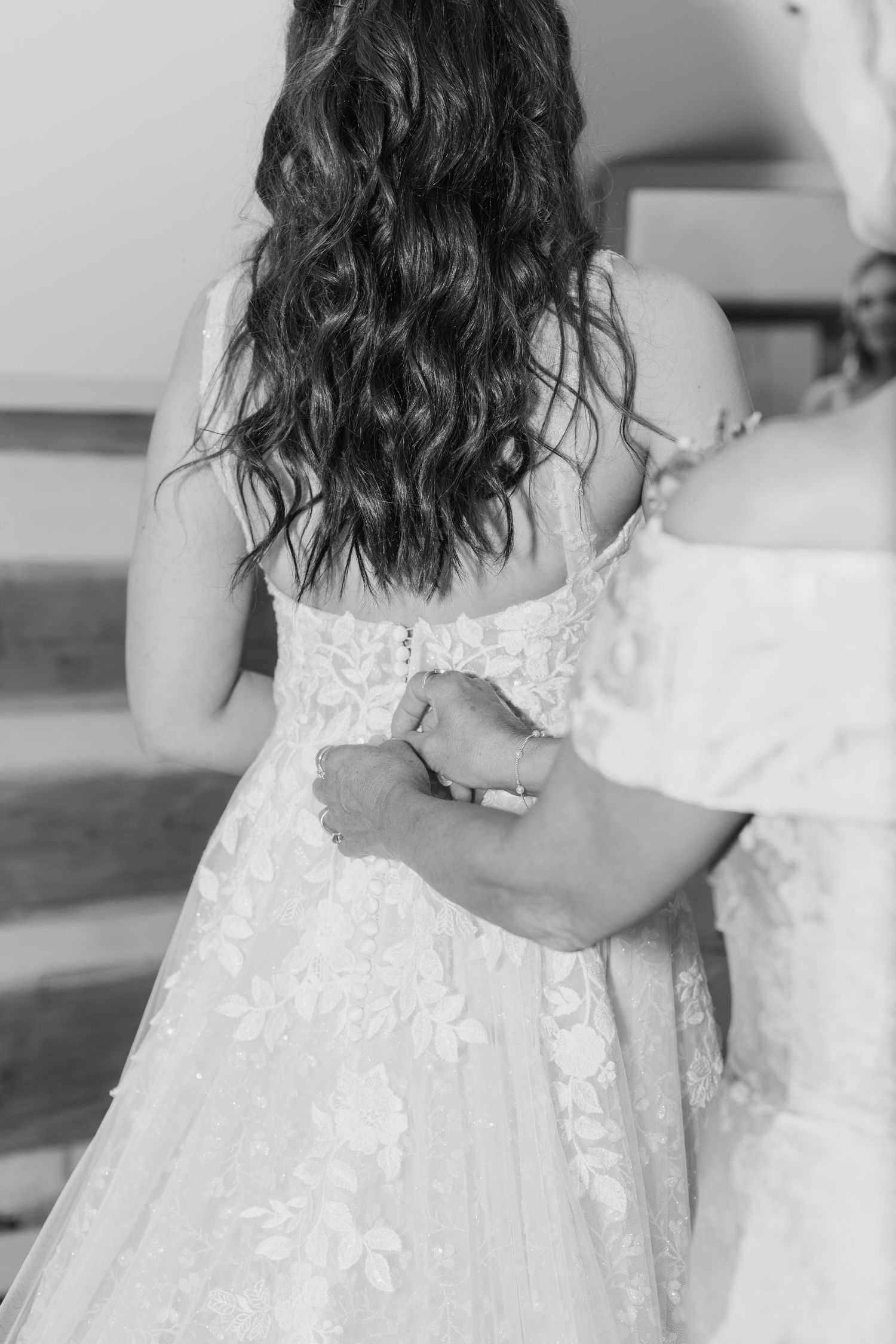 Black and white photo of bride in lace wedding gown being helped by bridesmaid, showing intricate back detail.