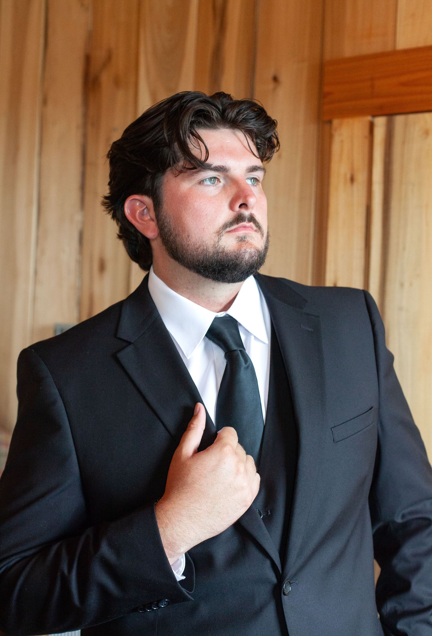 Young man in black suit adjusting tie, with dark curly hair and beard, standing against wooden wall backdrop.