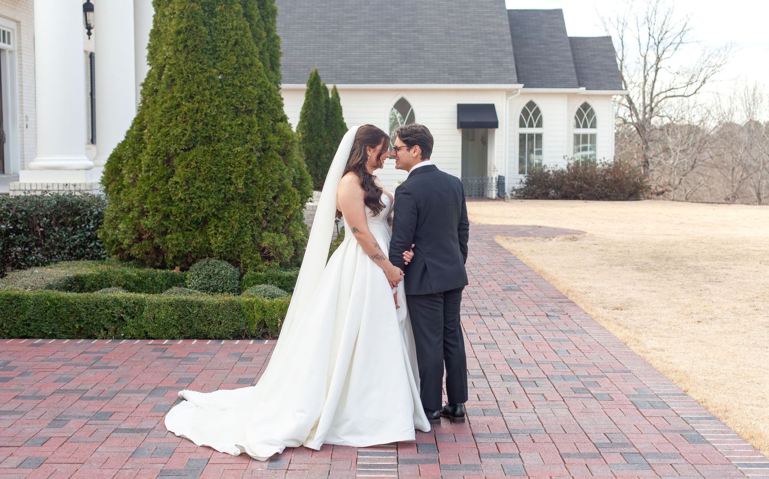 Bride and groom share an intimate moment on brick pathway outside elegant white wedding venue.