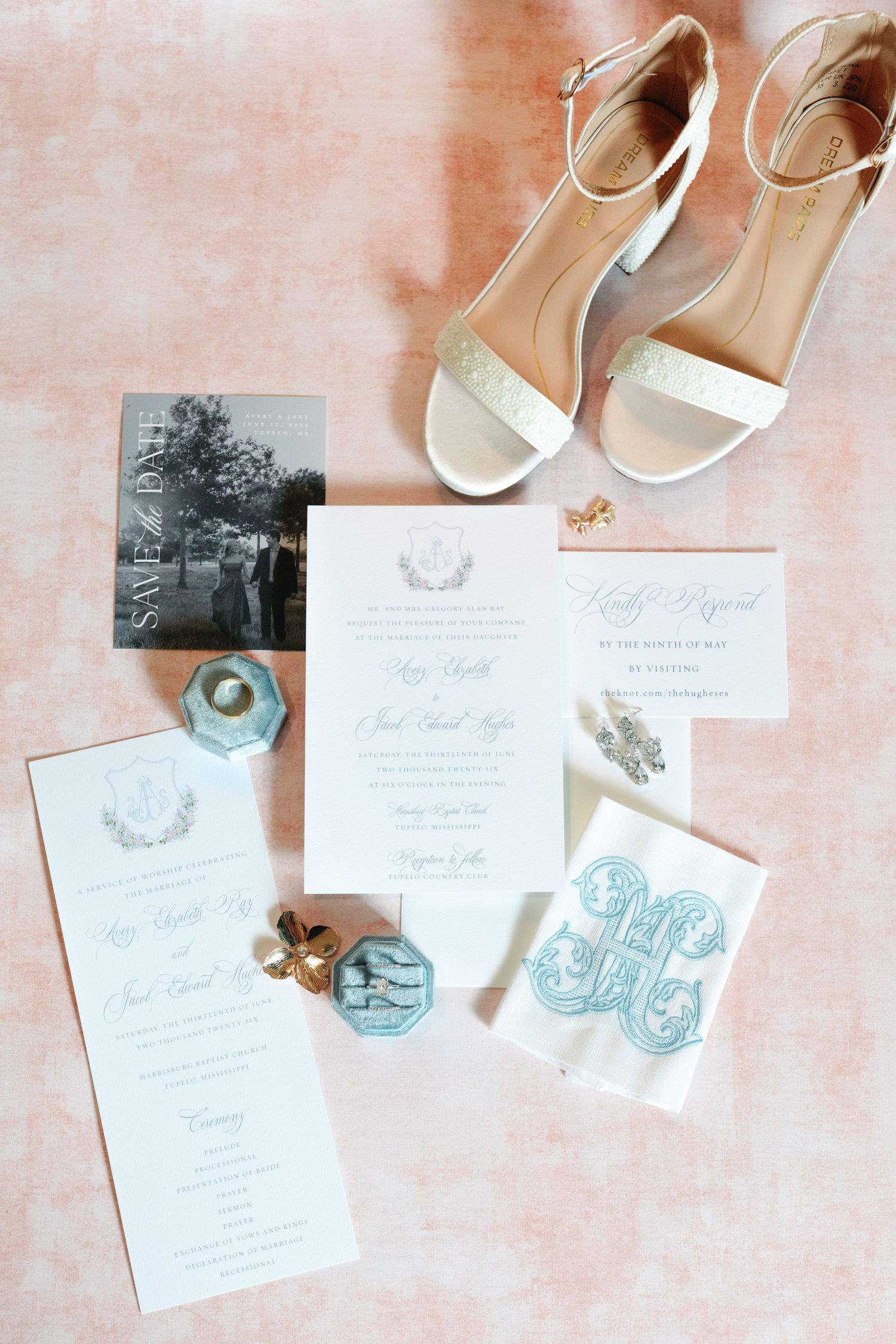 Elegant wedding flat lay with gold heels, blue monogram details, invitations, save the date, and jewelry on pink backdrop.