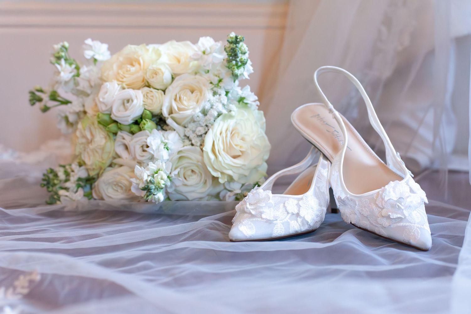 White bridal heels with lace detail beside an elegant white rose bouquet on soft gray fabric.