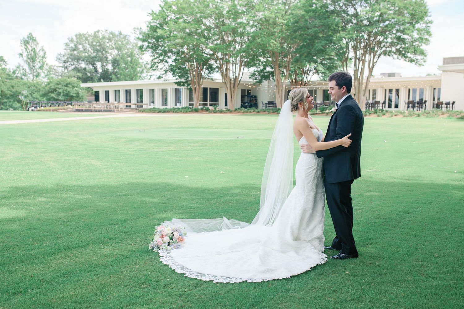 Bride and groom share a romantic moment on lush green lawn outside elegant wedding venue.