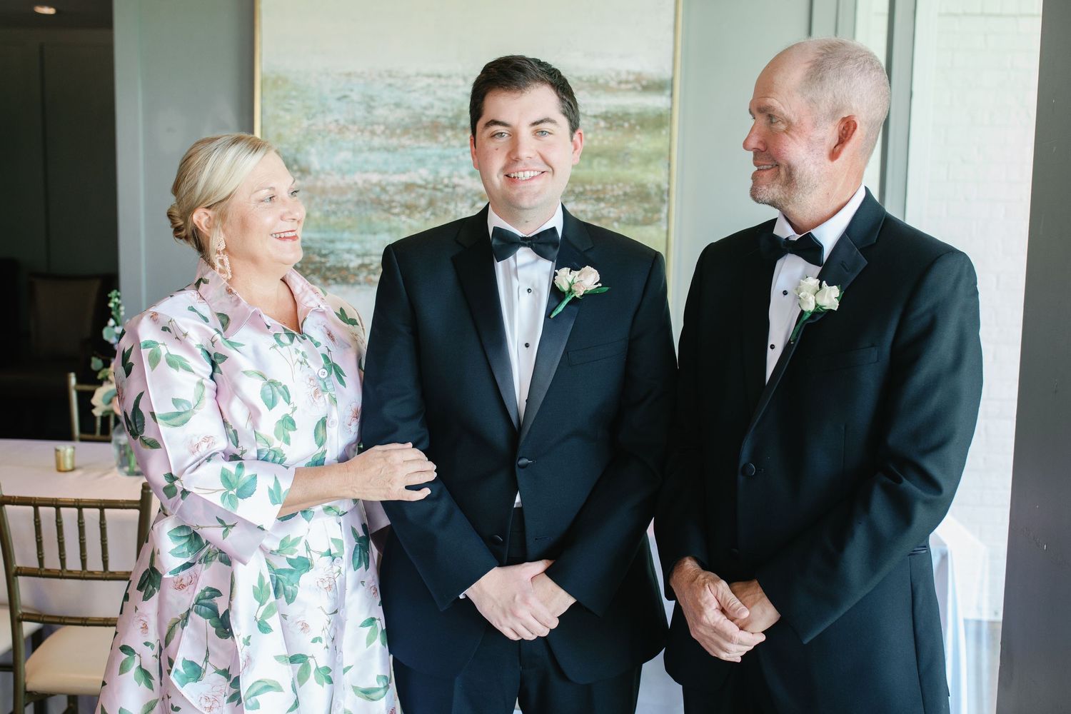 Groom in tuxedo smiling with parents at wedding, mother in floral dress, father in black suit.