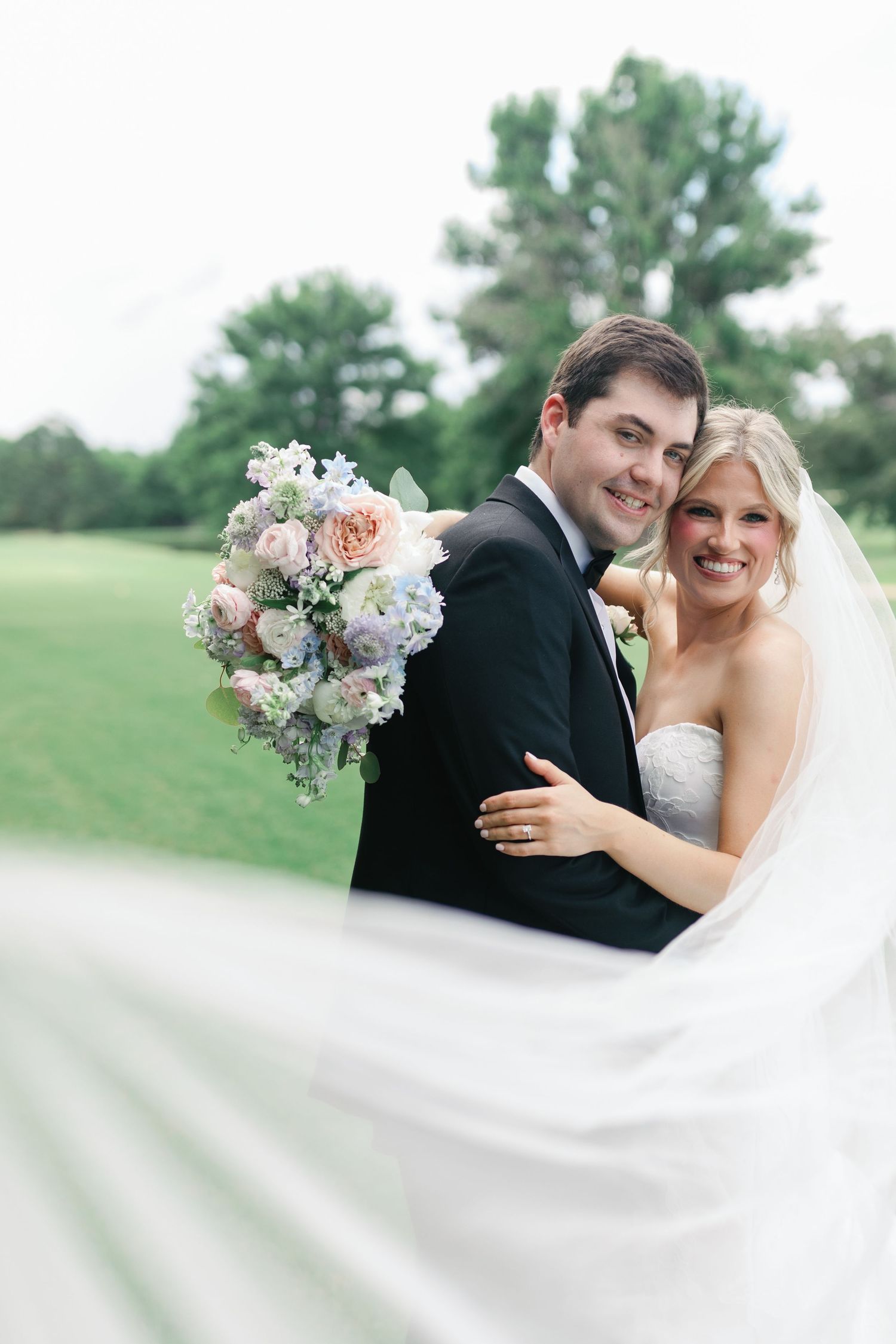 Smiling bride and groom embrace outdoors, her veil flowing, holding a pastel bouquet of roses and hydrangeas on their wedding day.