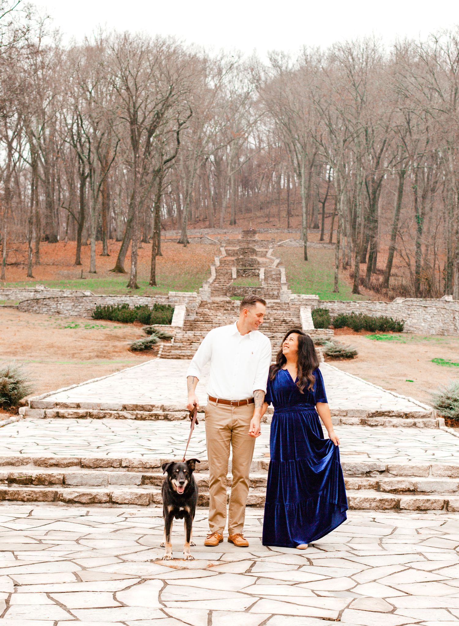 A couple in a blue dress and khaki attire share a romantic moment on a stone patio surrounded by winter trees.
