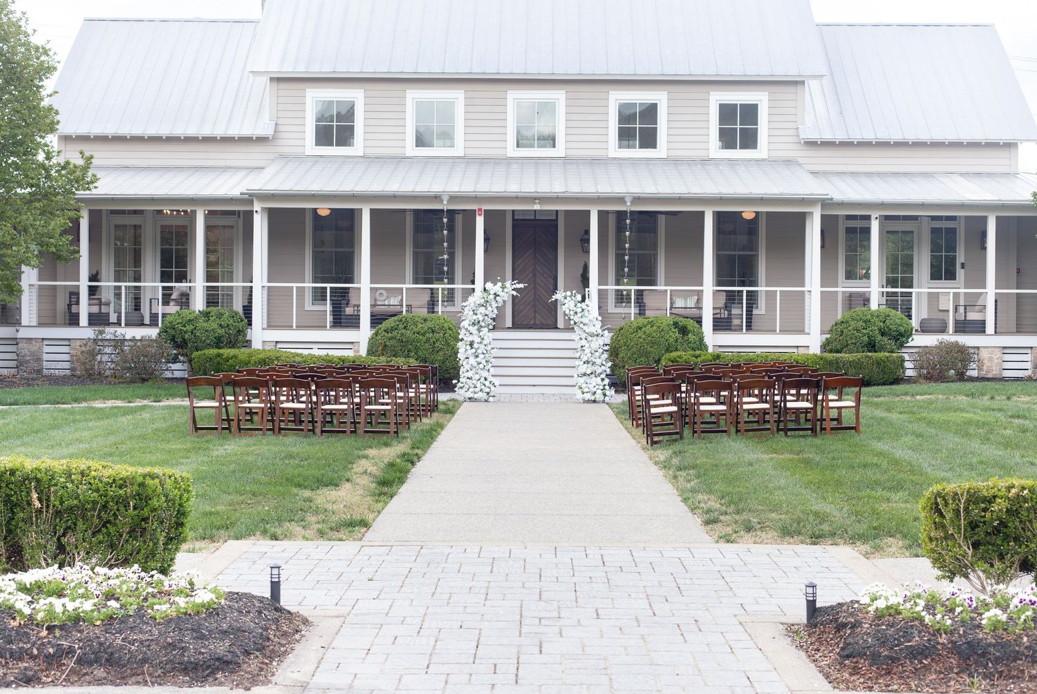Elegant farmhouse wedding venue with white arch, wooden chairs, manicured hedges, and stone pathway leading to wrap-around porch.