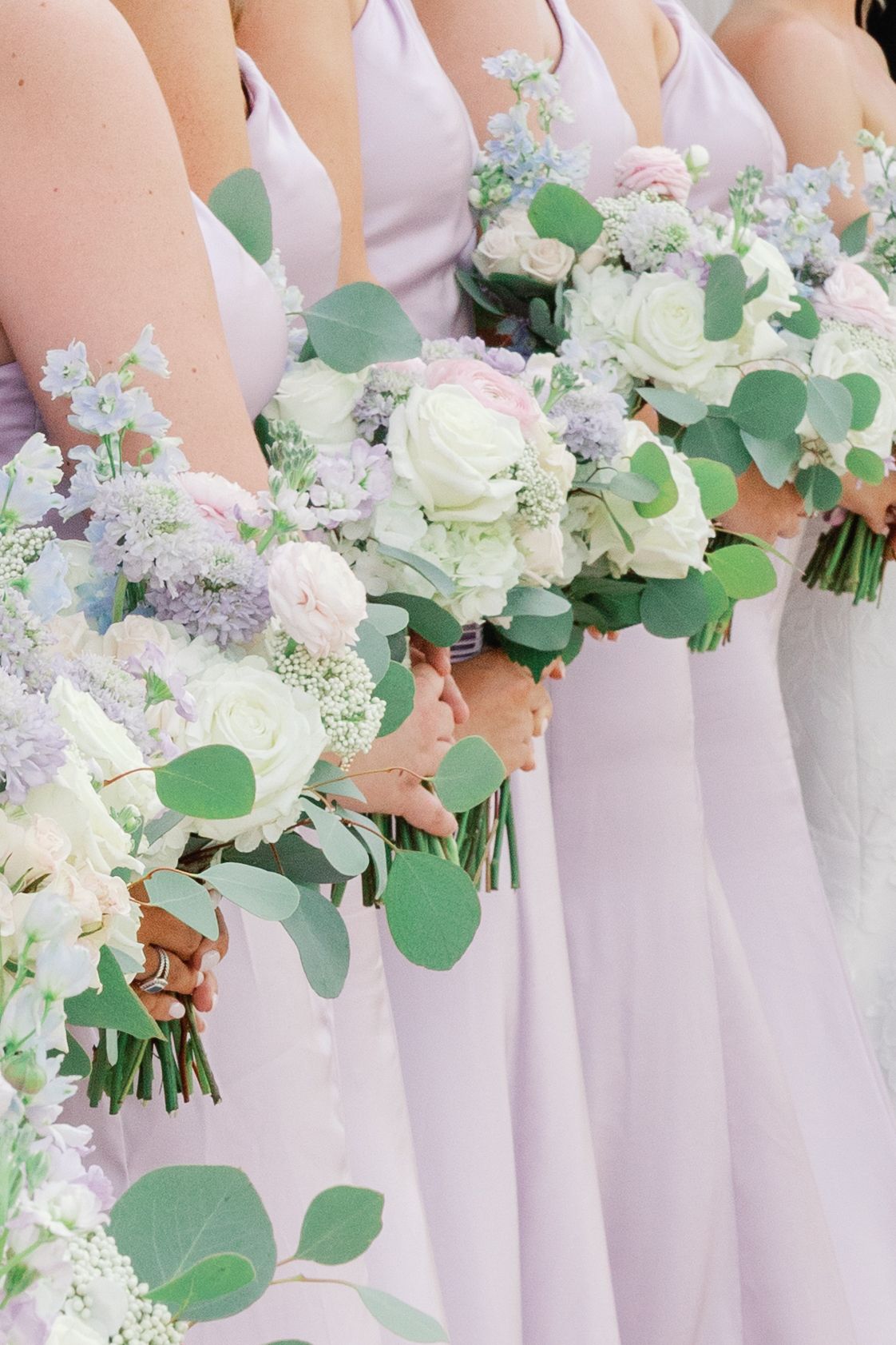 Bridesmaids in lavender gowns holding elegant bouquets of white roses, eucalyptus, and purple flowers at a wedding.