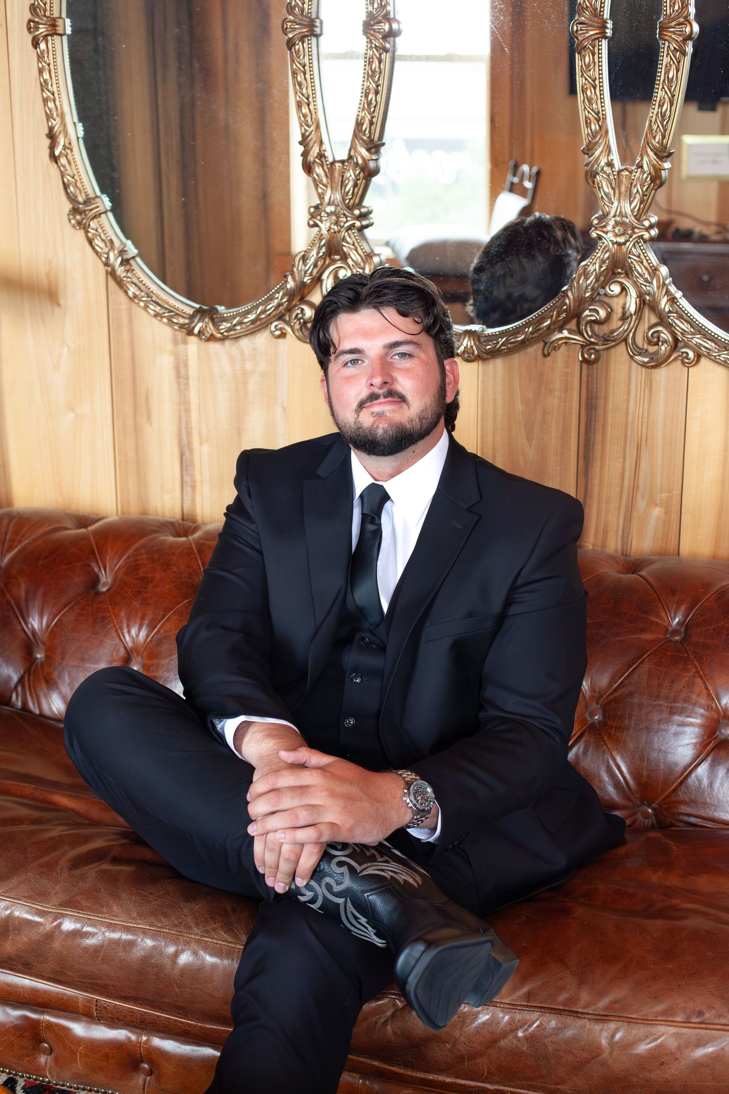 Man in black suit sitting on leather couch with ornate gold mirrors behind him.