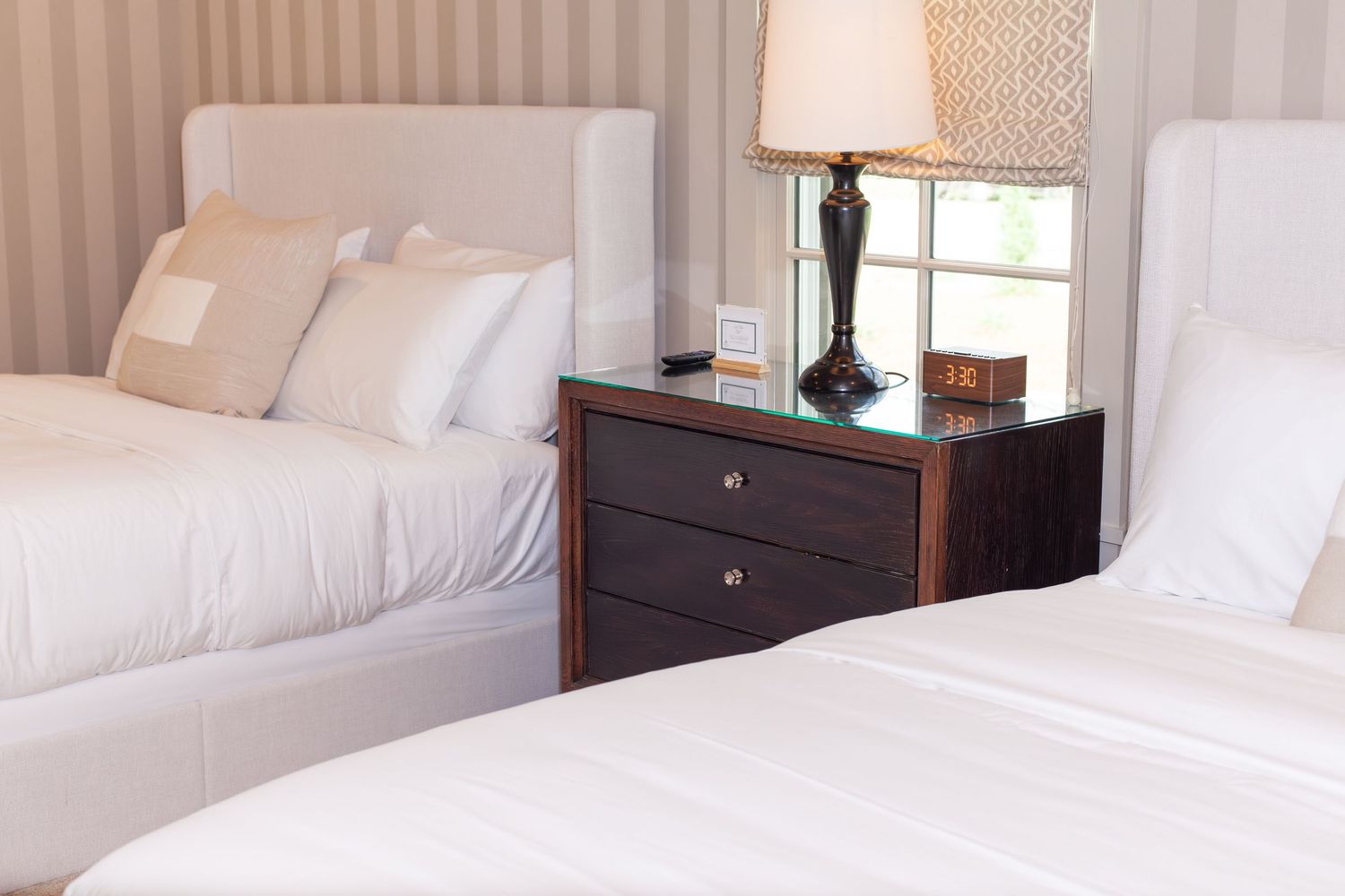 Two white hotel beds with a dark wood nightstand and lamp between them in an elegant striped room.
