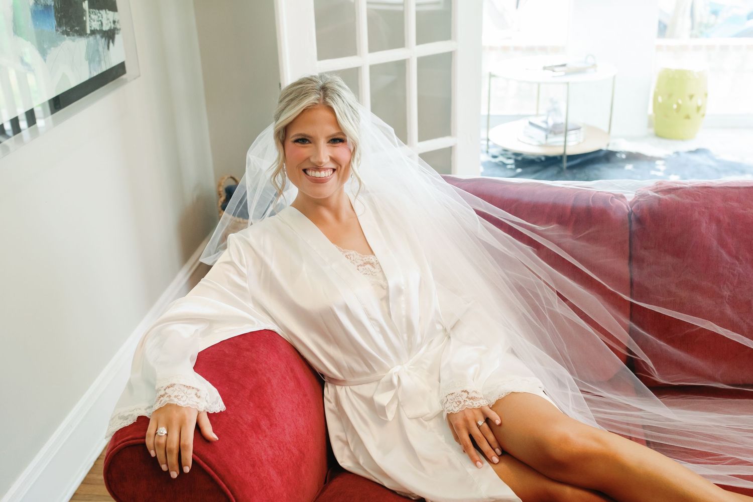 Smiling bride in white robe and veil sitting on red sofa near window during wedding preparation.