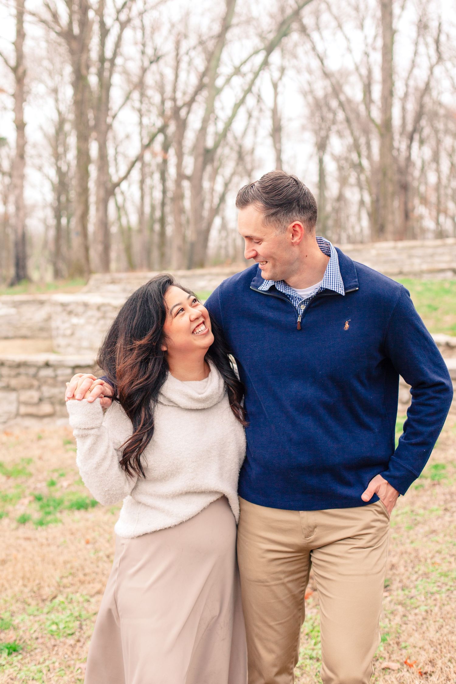 A couple shares joyful laughter while posing together against a stone wall in an autumn forest setting.