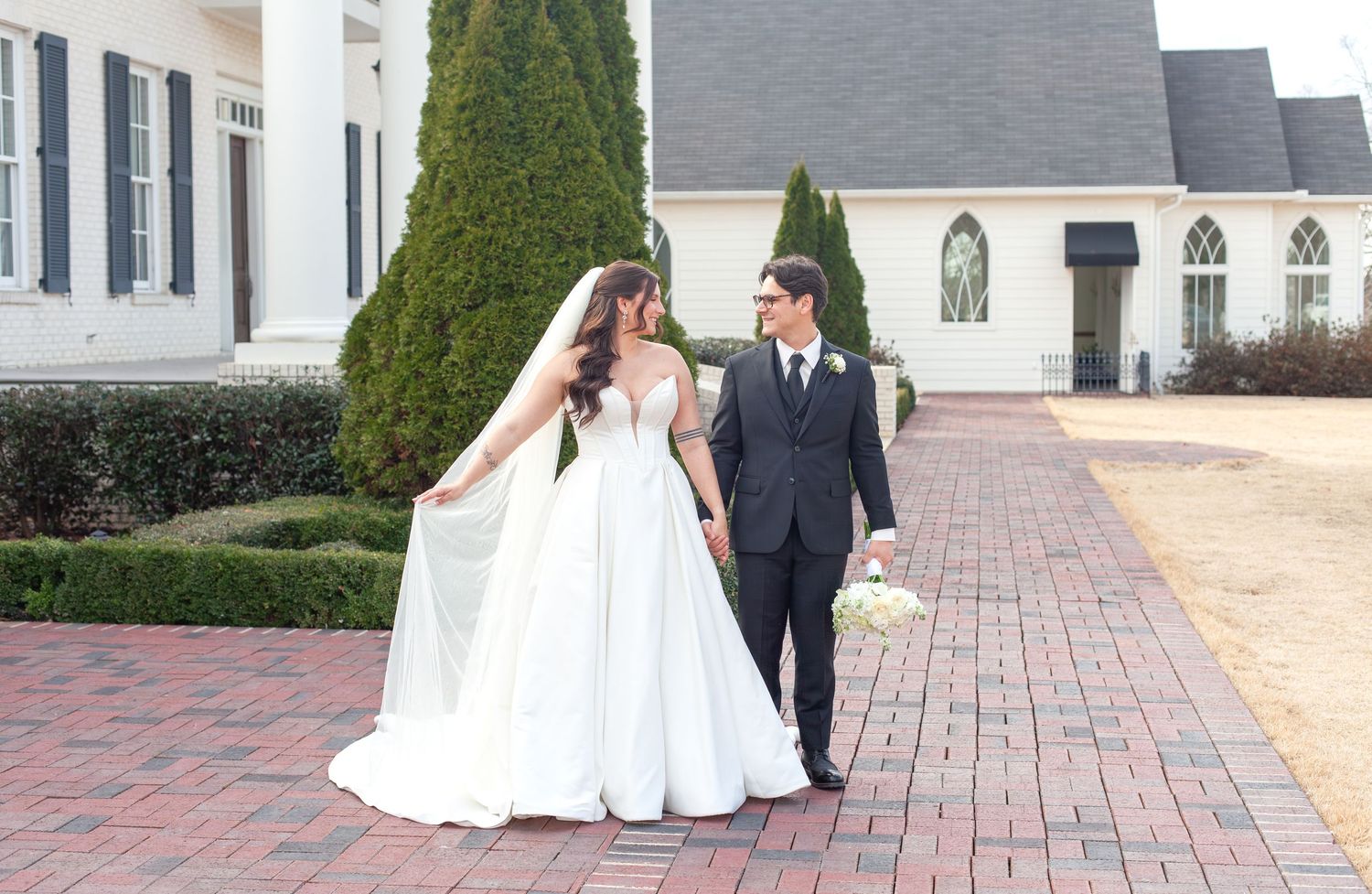 Bride and groom walking hand in hand on a brick pathway outside a white wedding venue with manicured hedges.