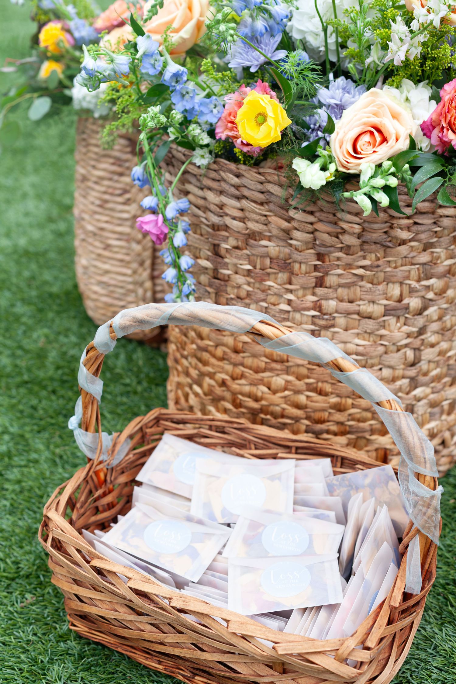 Wicker basket filled with envelopes beside a large floral arrangement in a woven basket on green grass.
