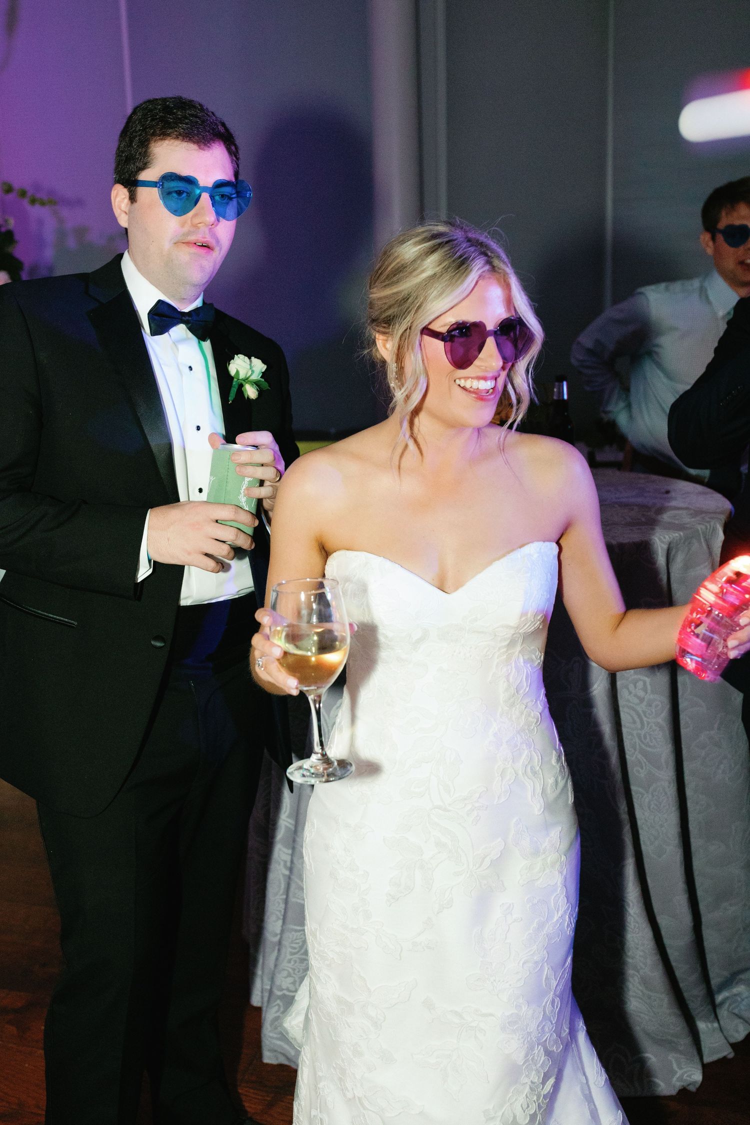 Bride and groom wearing heart-shaped sunglasses, holding drinks and smiling at wedding reception.