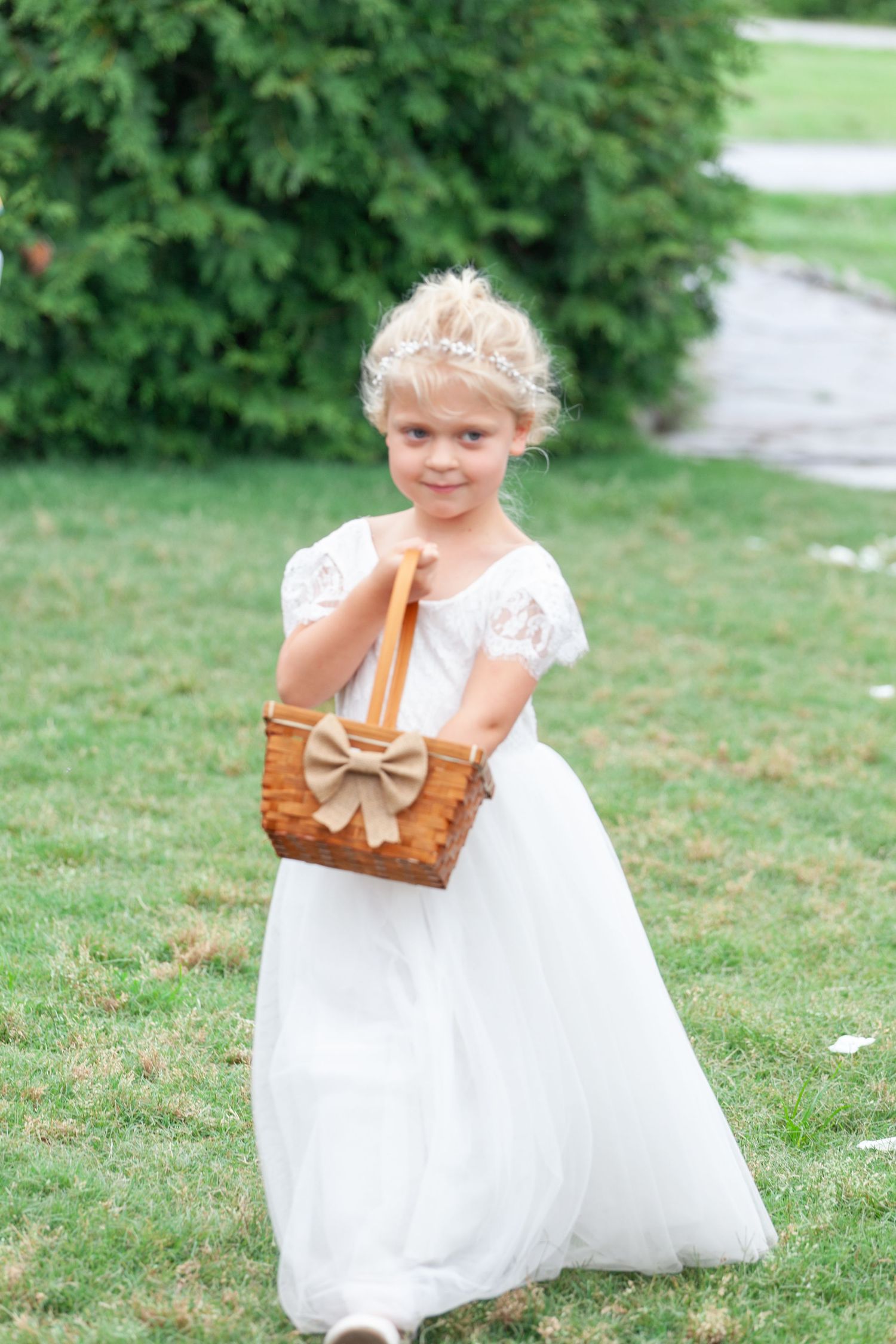 Adorable flower girl in white dress holding a wicker basket with burlap bow at an outdoor wedding ceremony.
