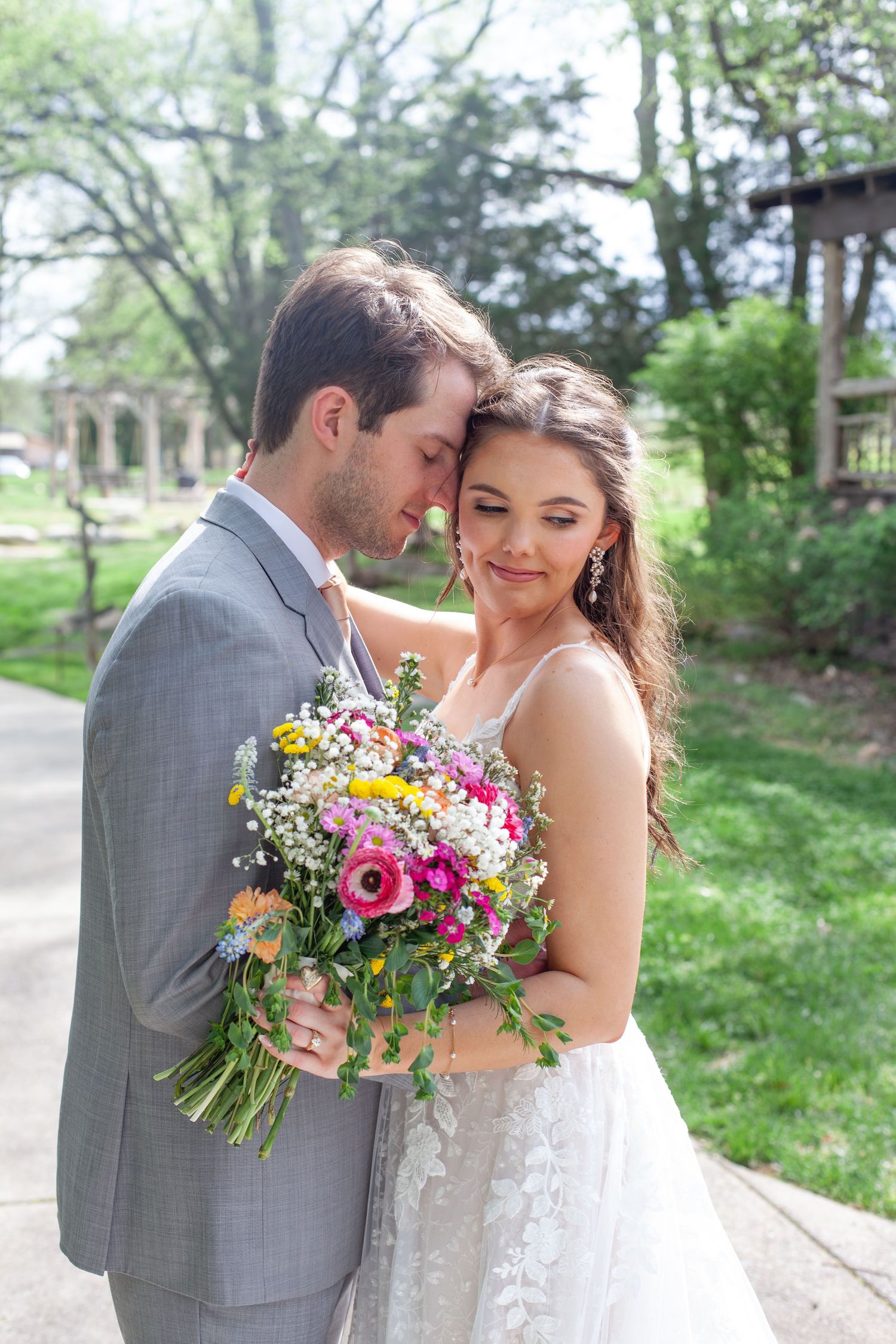 A bride holding a colorful wildflower bouquet leans into her groom in a gray suit outdoors.