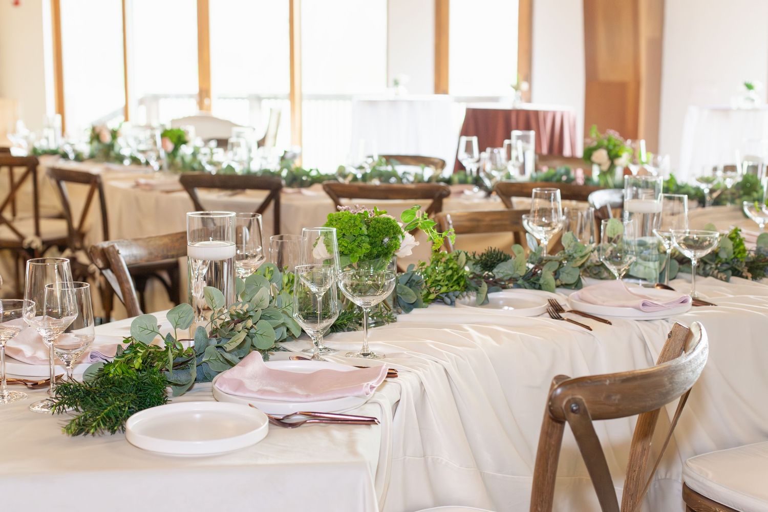 Elegant wedding reception table with greenery garland runner, crystal glasses, and pink napkins on white linen.