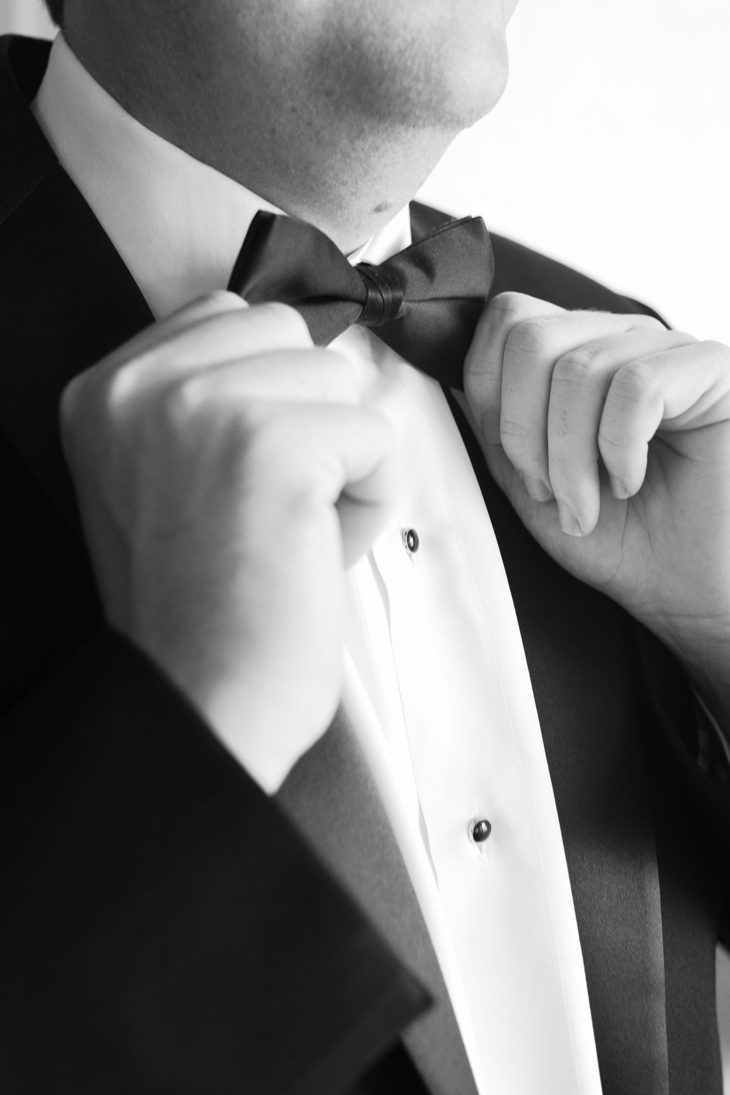 Black and white photo of a man in a tuxedo adjusting his bow tie with both hands.