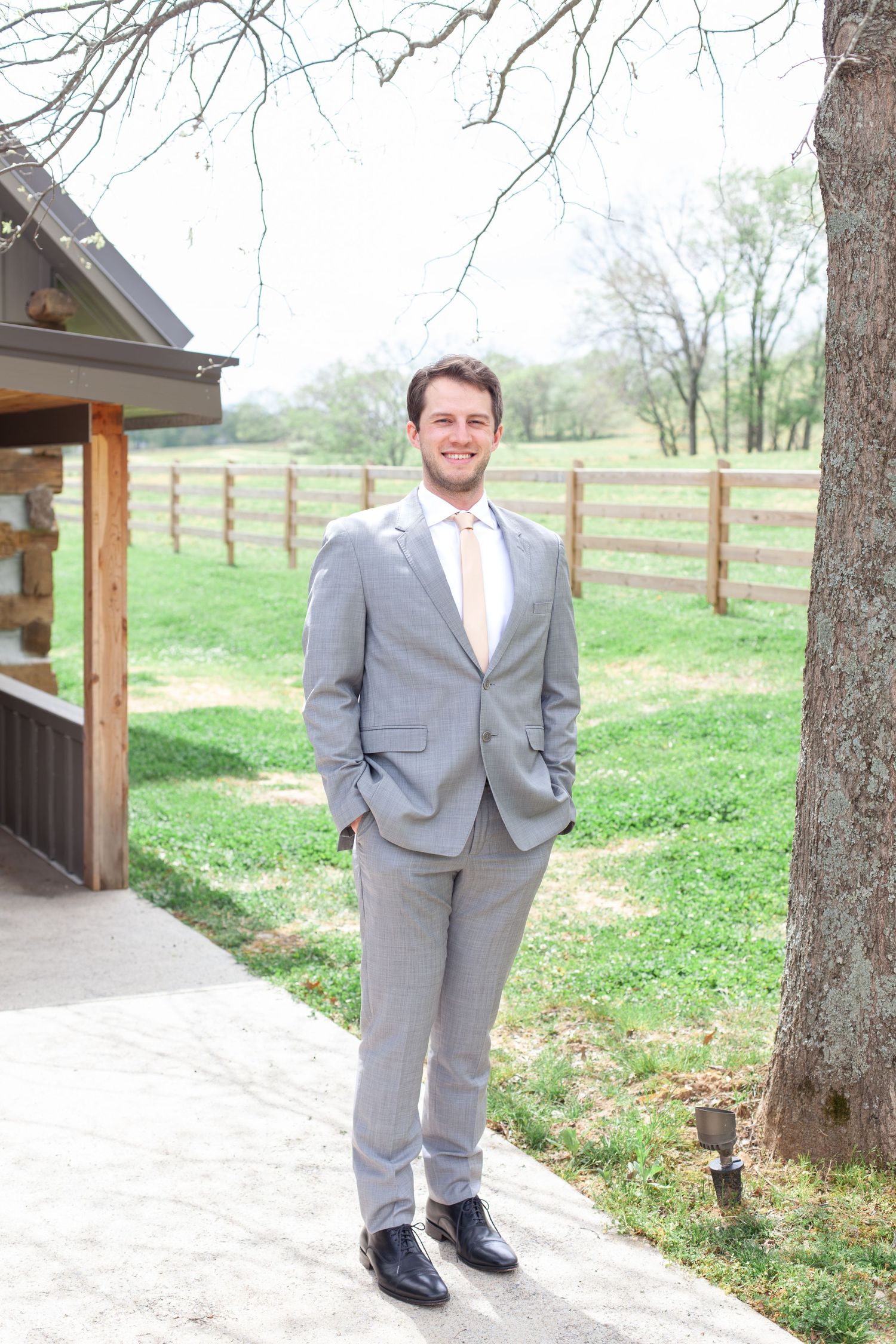 Smiling groom in gray suit and blush tie stands outdoors near rustic fence and barn on wedding day.