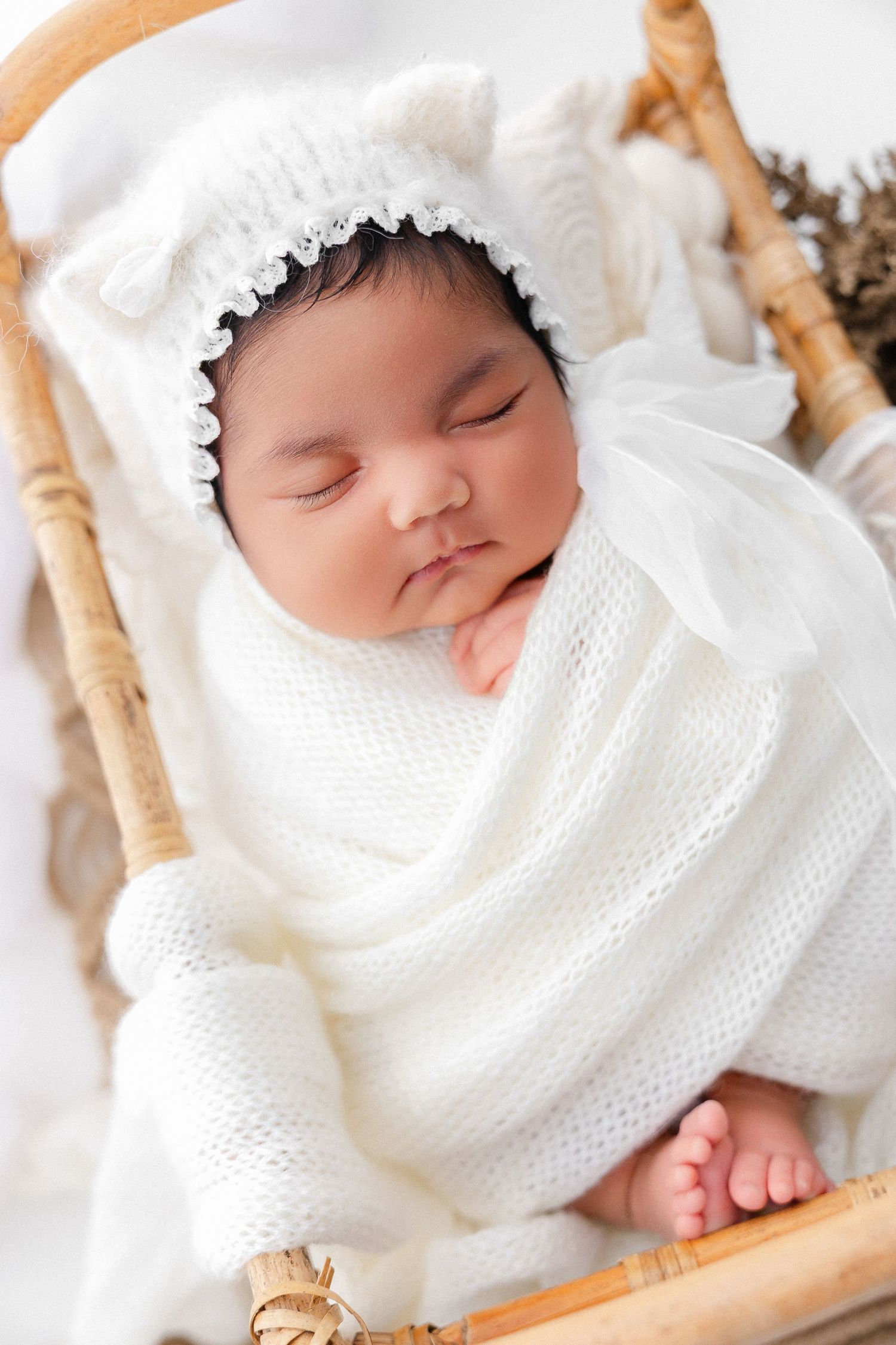 Sleeping baby wrapped in white knit blanket resting in wooden basket with delicate details.