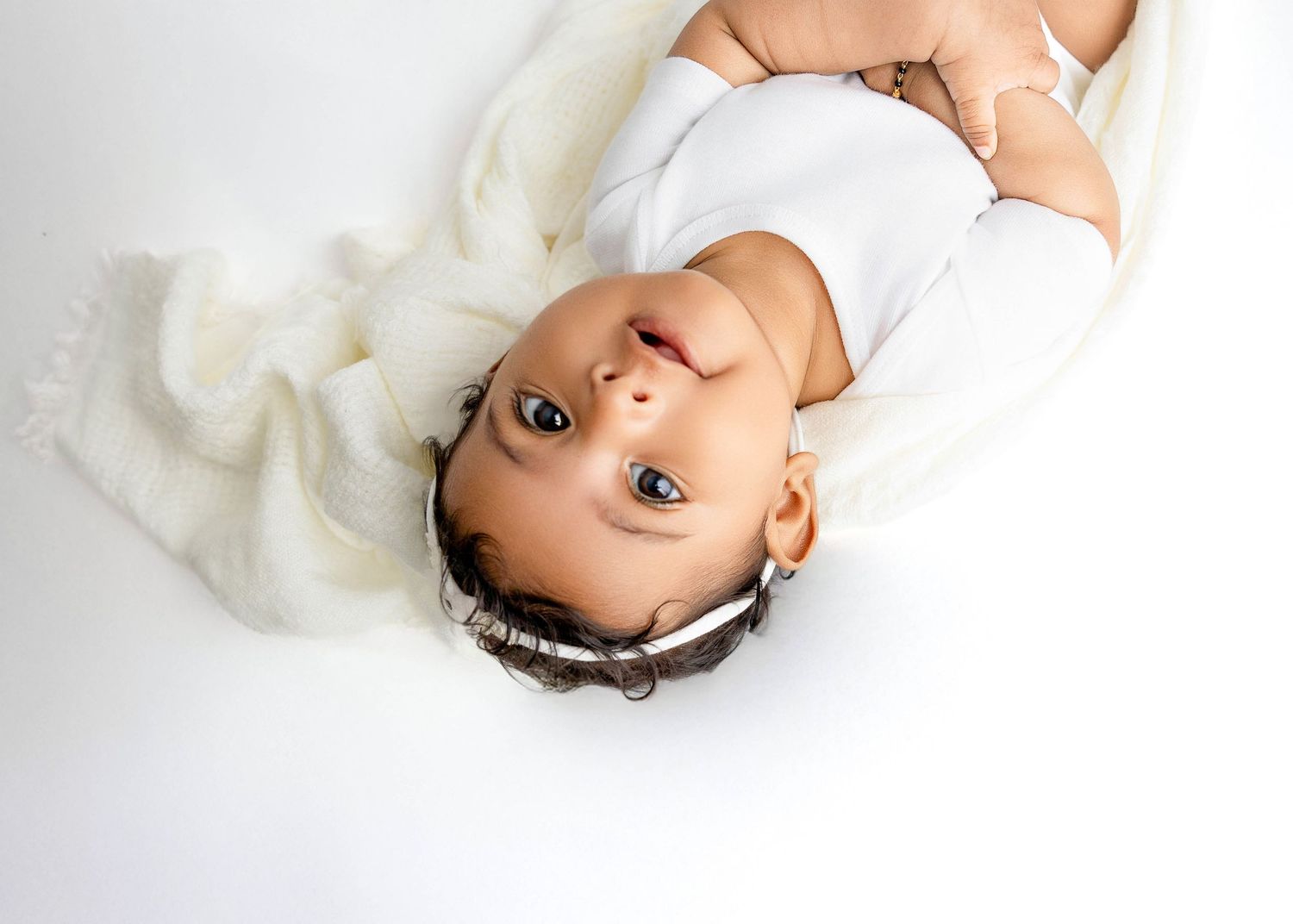 Baby lying on white fabric with hands reaching up during a photo session.