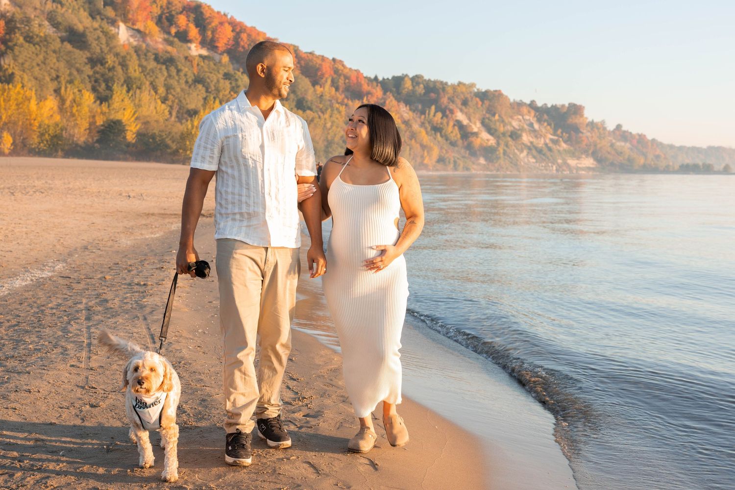 Sunset beach maternity photos with couple walking along shoreline in white and beige outfits.