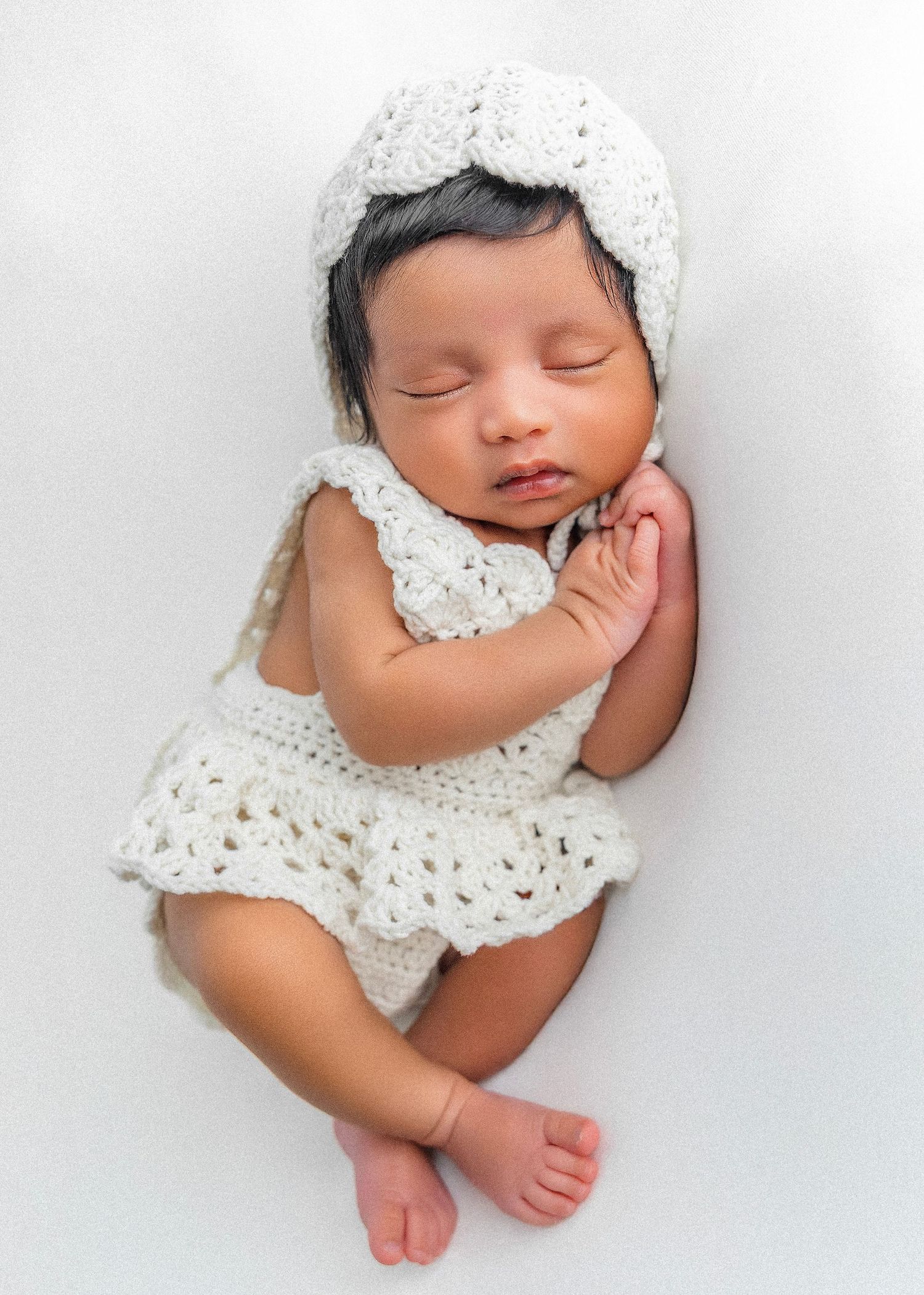 Sleeping newborn wearing white lace bonnet and dress against soft white background.