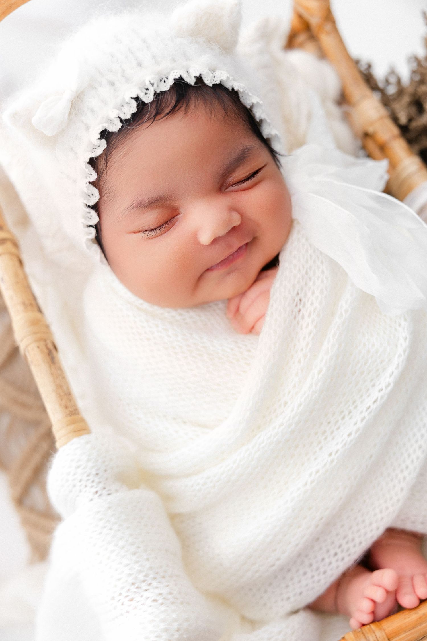 Sleeping baby wearing white knit sweater and lace bonnet on woven surface.