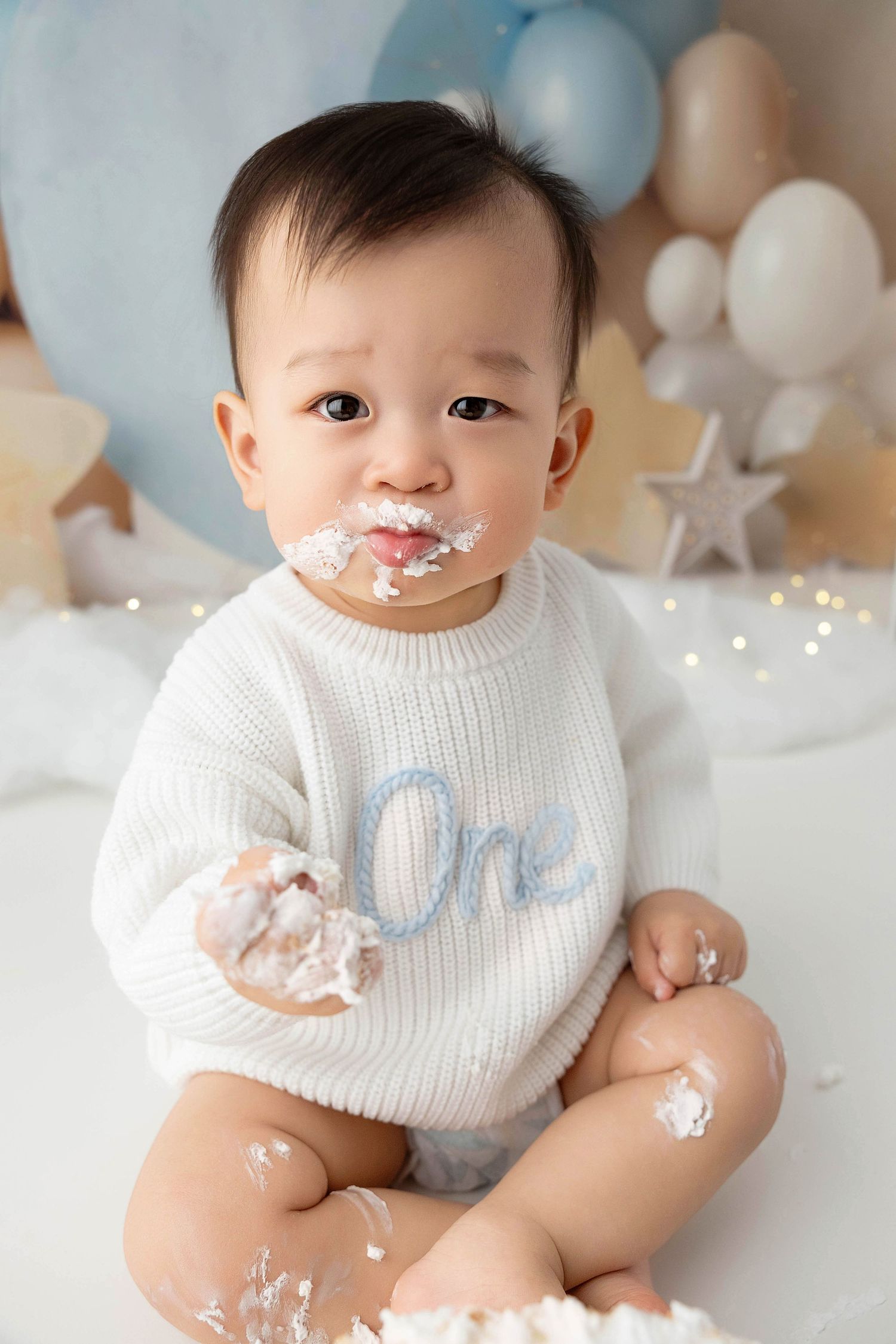 Baby wearing white knit sweater during cake smash photoshoot with decorations.