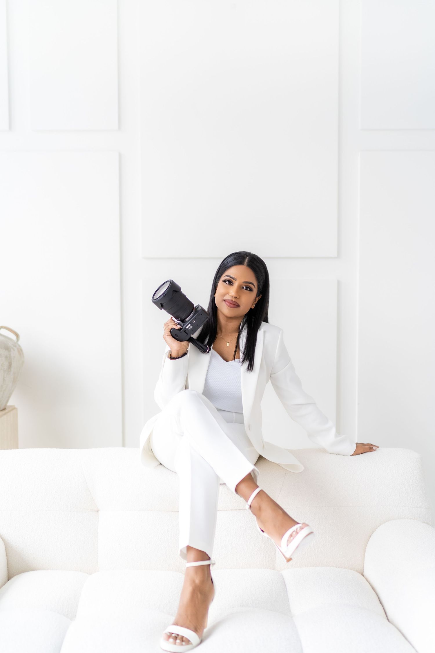 Female photographer in white blazer holding camera against white studio backdrop.