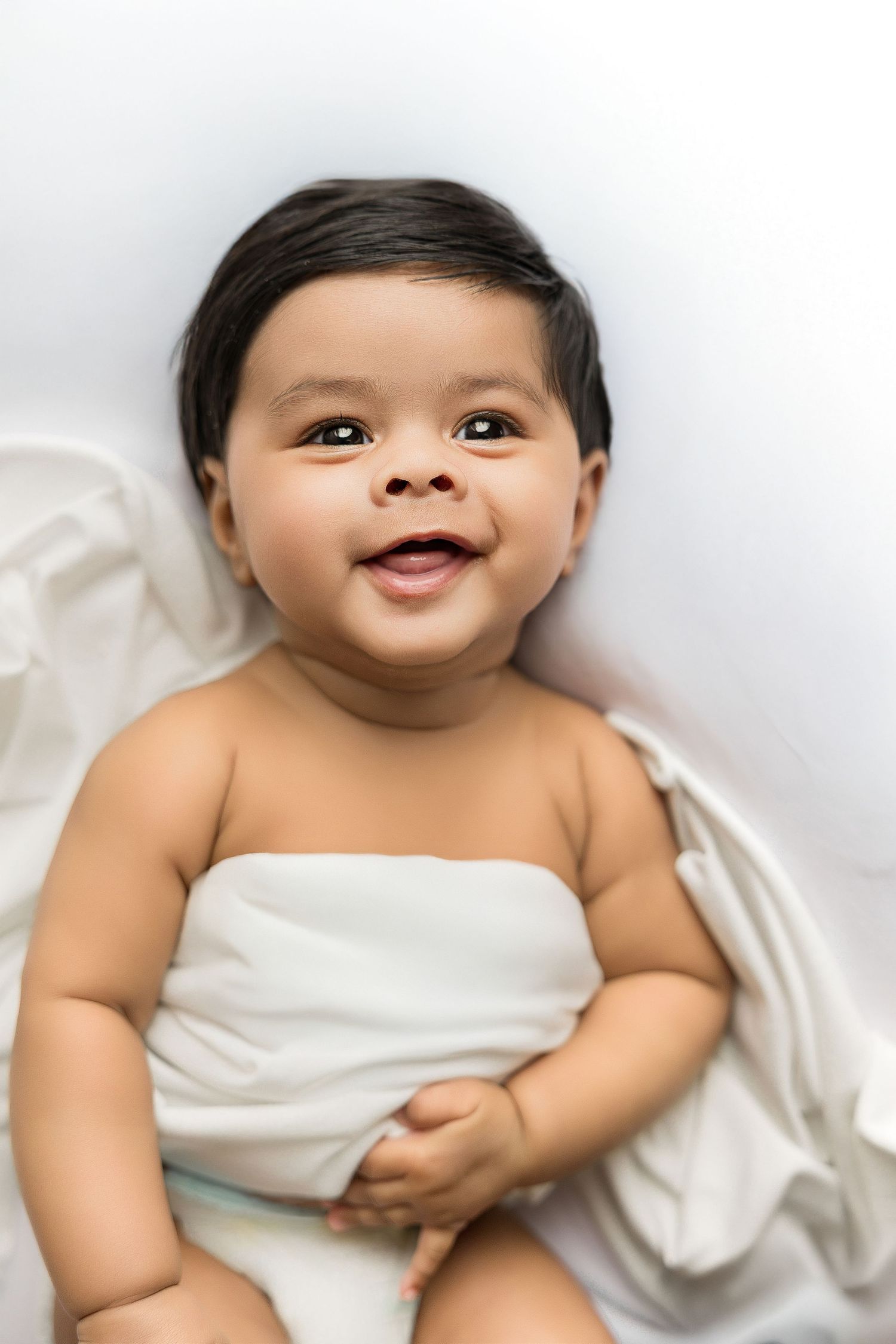 A happy baby wrapped in white fabric smiles brightly while lying down on a light background.
