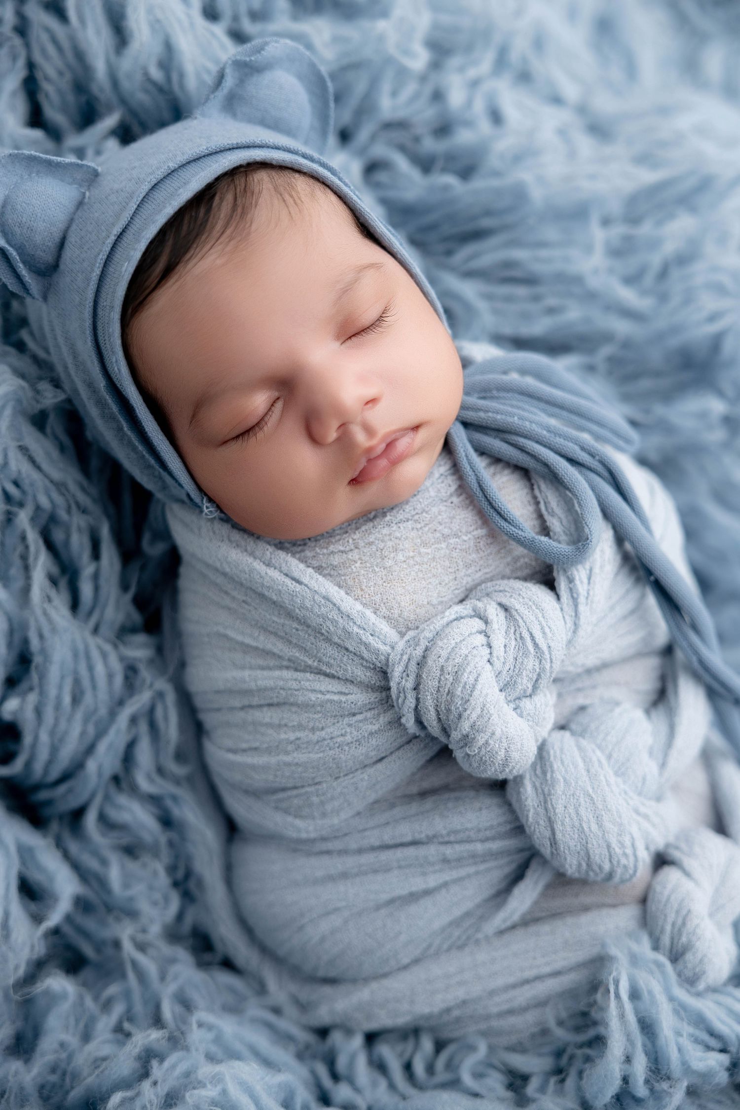 Peaceful newborn swaddled in soft blue wrap on textured blue backdrop in professional photo series.