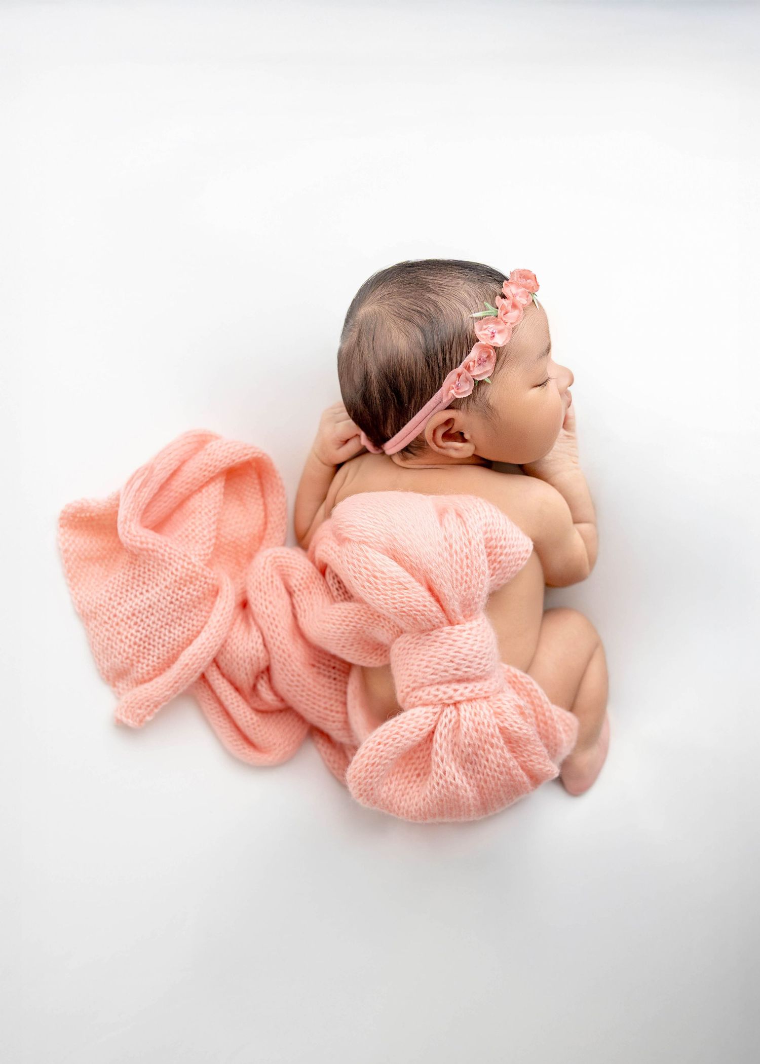 Newborn baby wearing soft pink knit wrap lying peacefully on white backdrop in artistic portrait series.