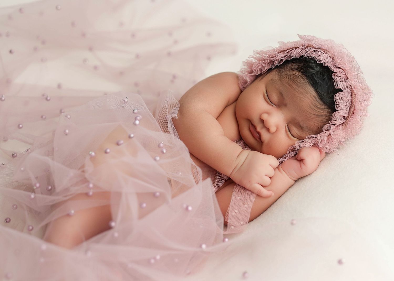 Newborn baby sleeping under pink tulle fabric in dreamy portrait setting.