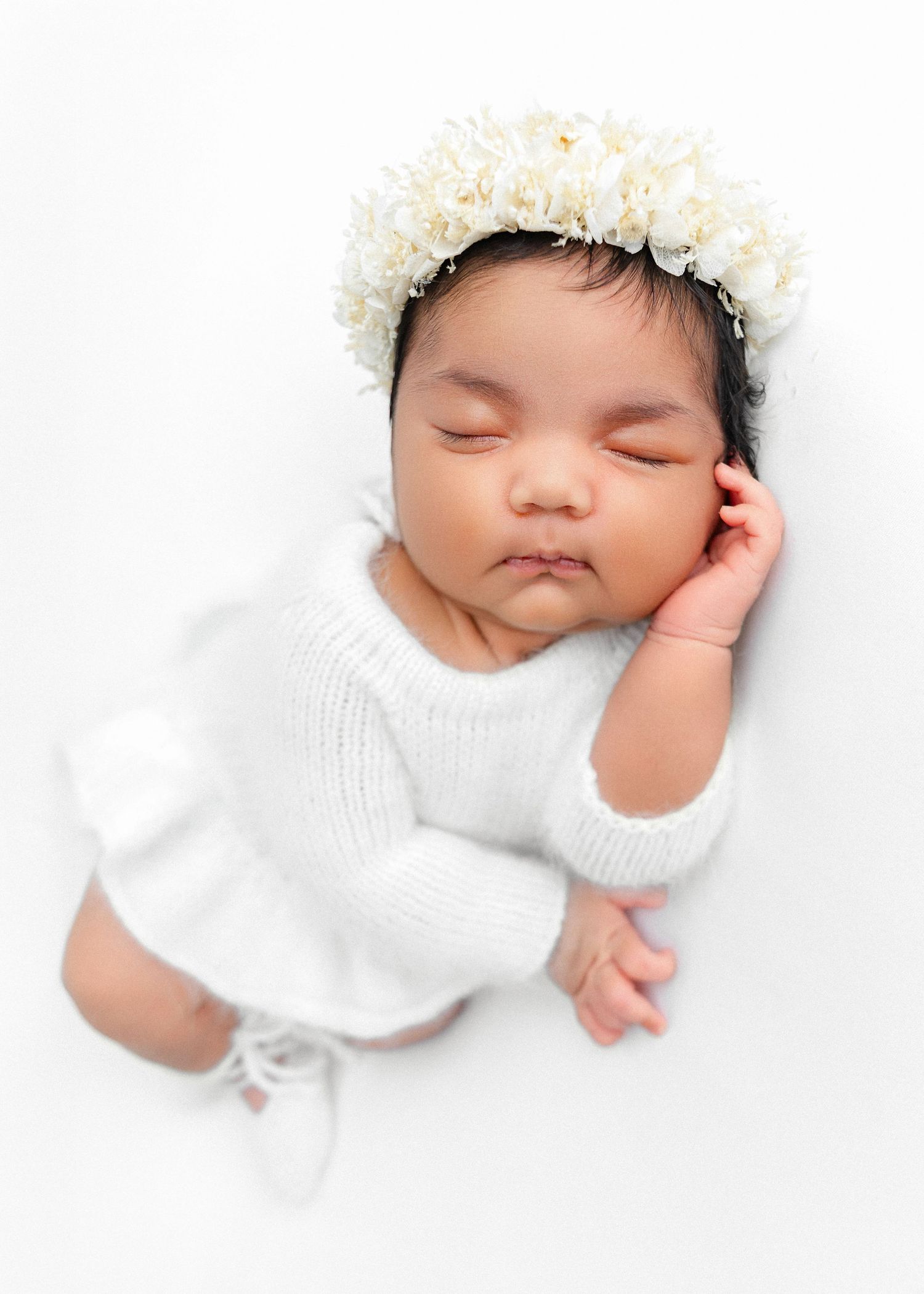 Sleeping newborn in white outfit with floral headpiece against white backdrop.