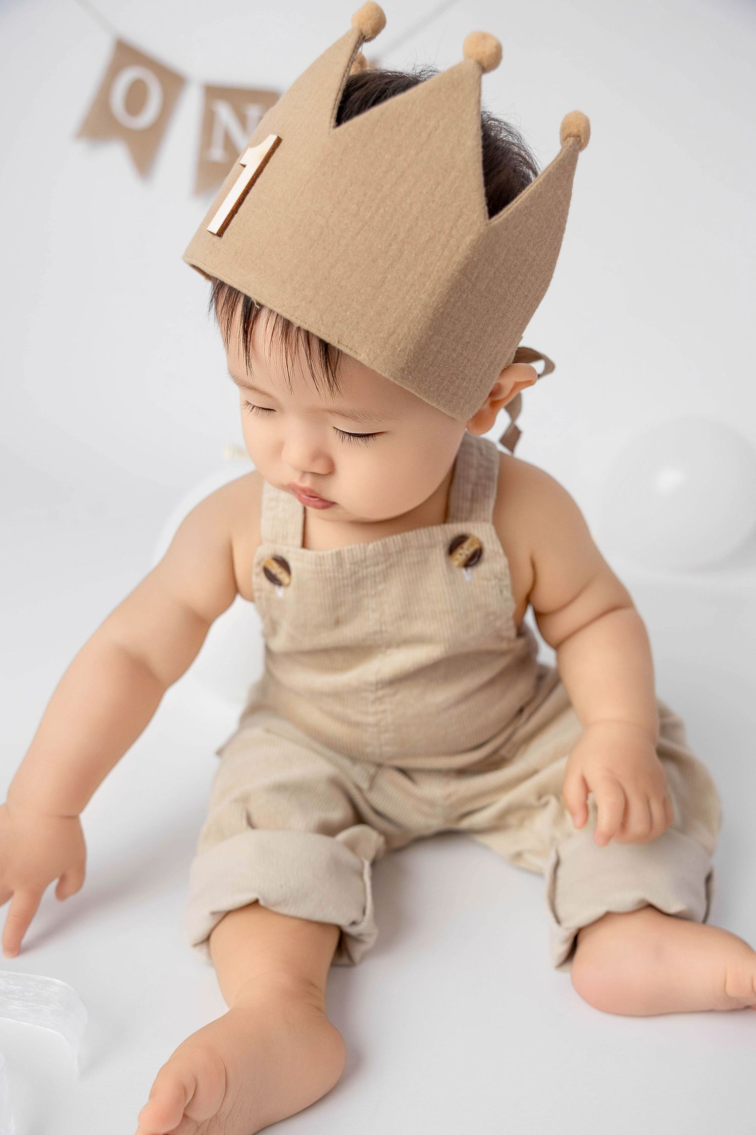 Baby wearing beige overalls and cardboard crown in minimalist studio setting.