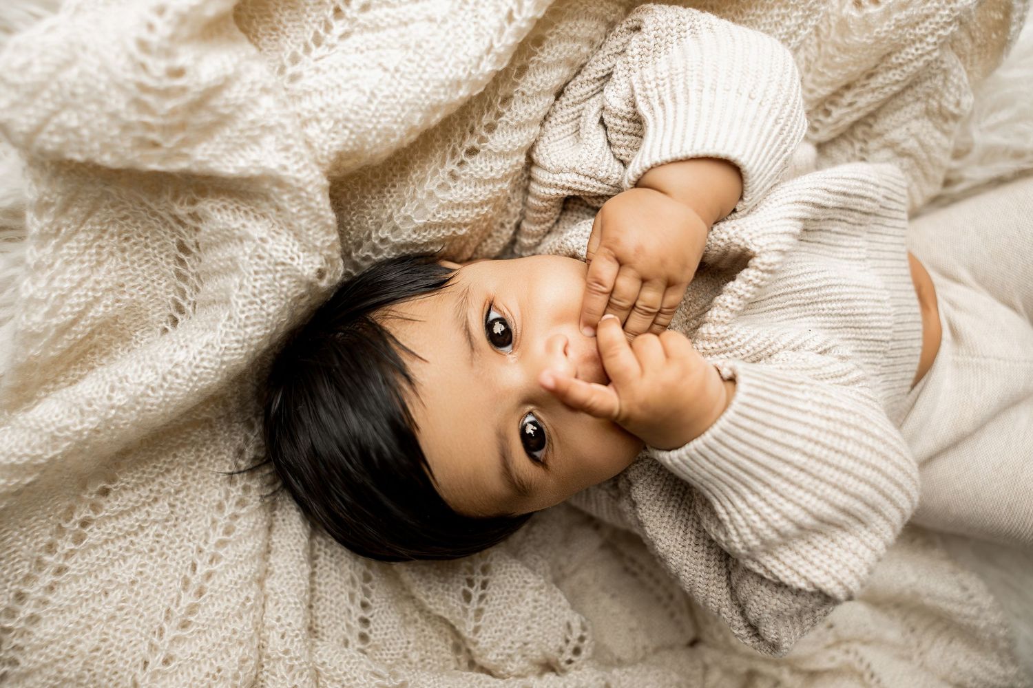 Baby wearing cozy cream sweater lying on white textured surface in photo series.