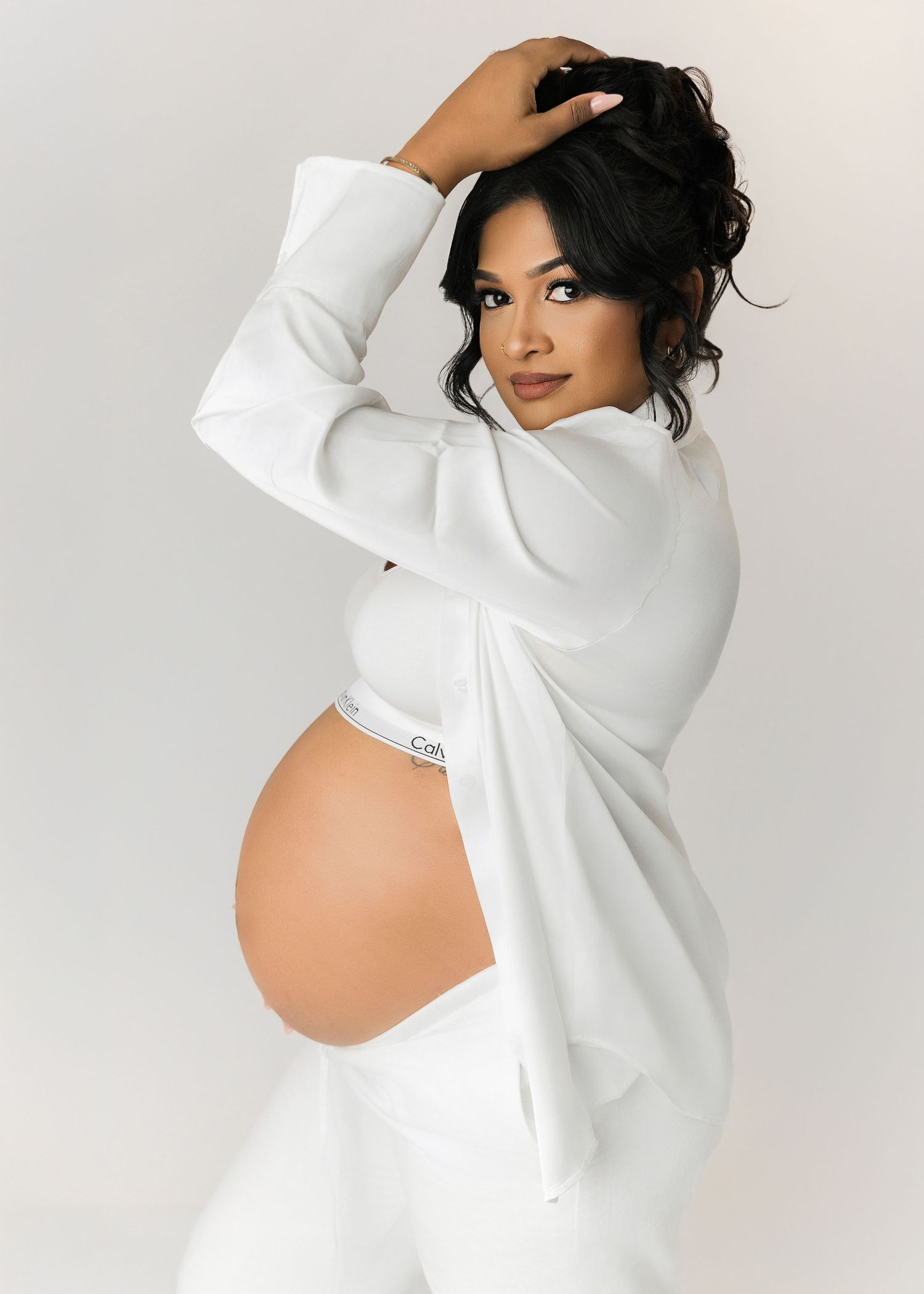 Elegant maternity portraits in white sweater showing pregnancy silhouette against light backdrop.