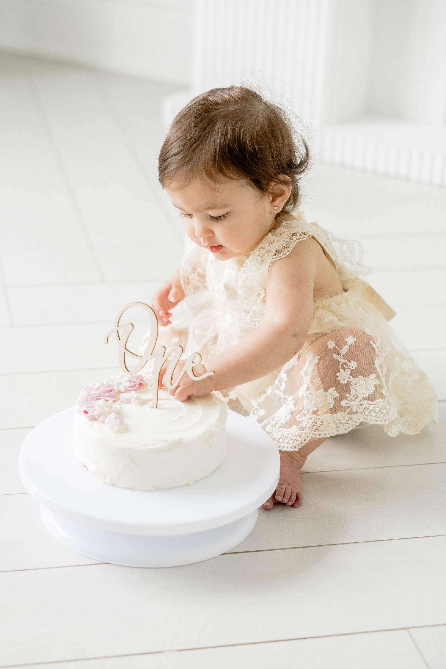 Baby in lace dress leaning over to touch white cake during photoshoot.