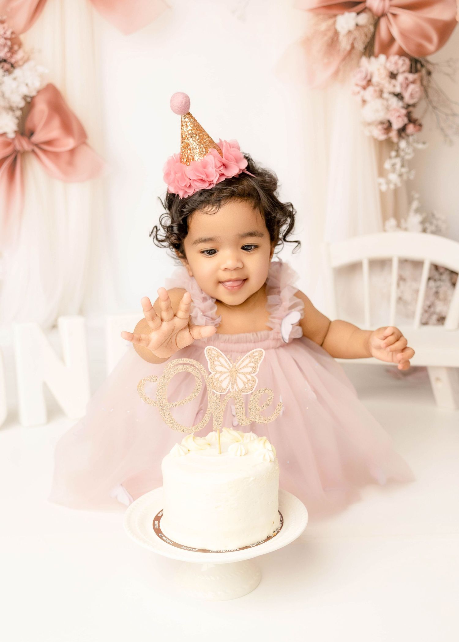 Baby in pink dress sitting with white birthday cake against floral backdrop.