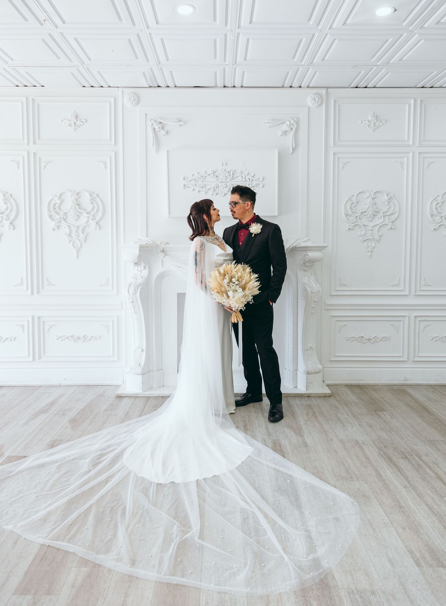 Couple in wedding attire standing by a white fireplace in an elegant room.