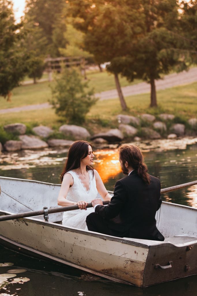 A couple in wedding attire sits in a rowboat during sunset on a peaceful lake.