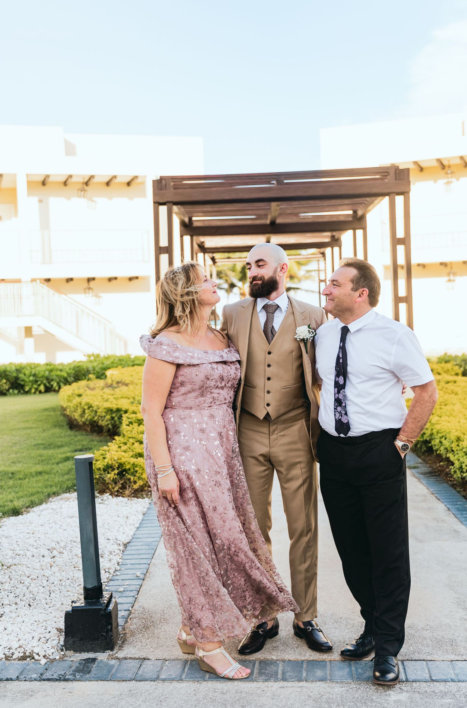 Wedding party members in pink dresses and tan suits pose together on a paved walkway at an outdoor venue.
