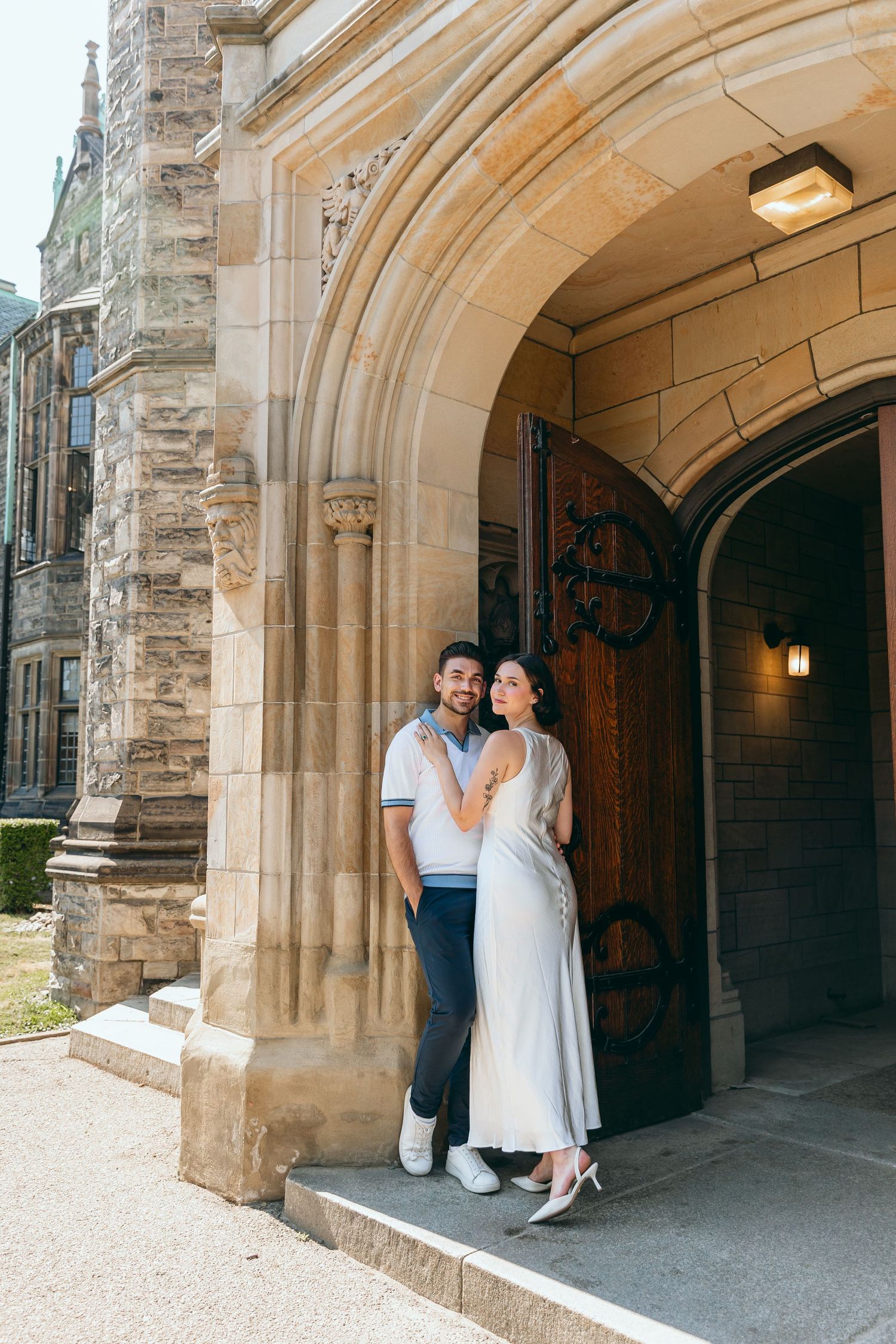 Couple poses beneath ornate stone archway entrance of historic building.