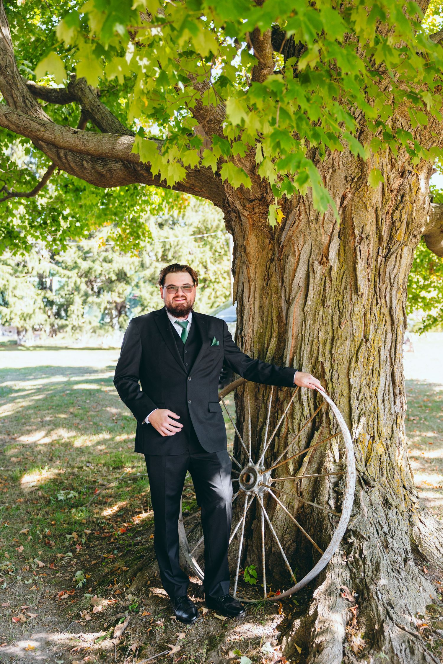 A rustic outdoor scene with a vintage wagon wheel leaning against a tree trunk in dappled sunlight.
