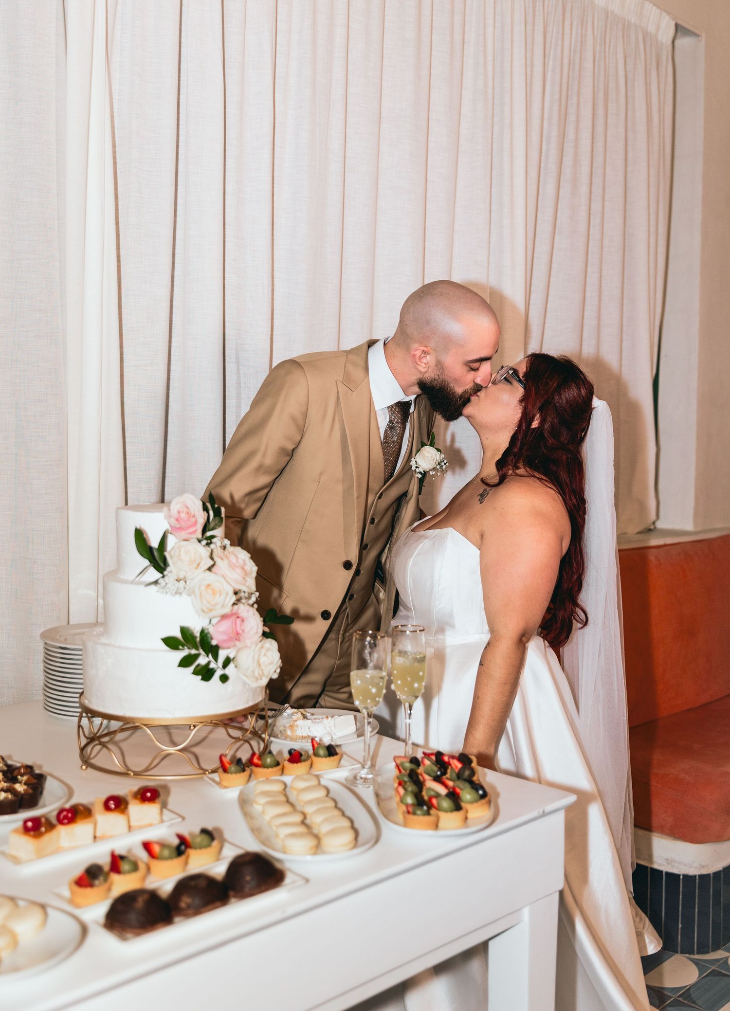 A couple shares a kiss beside their wedding dessert table with cake and appetizers.
