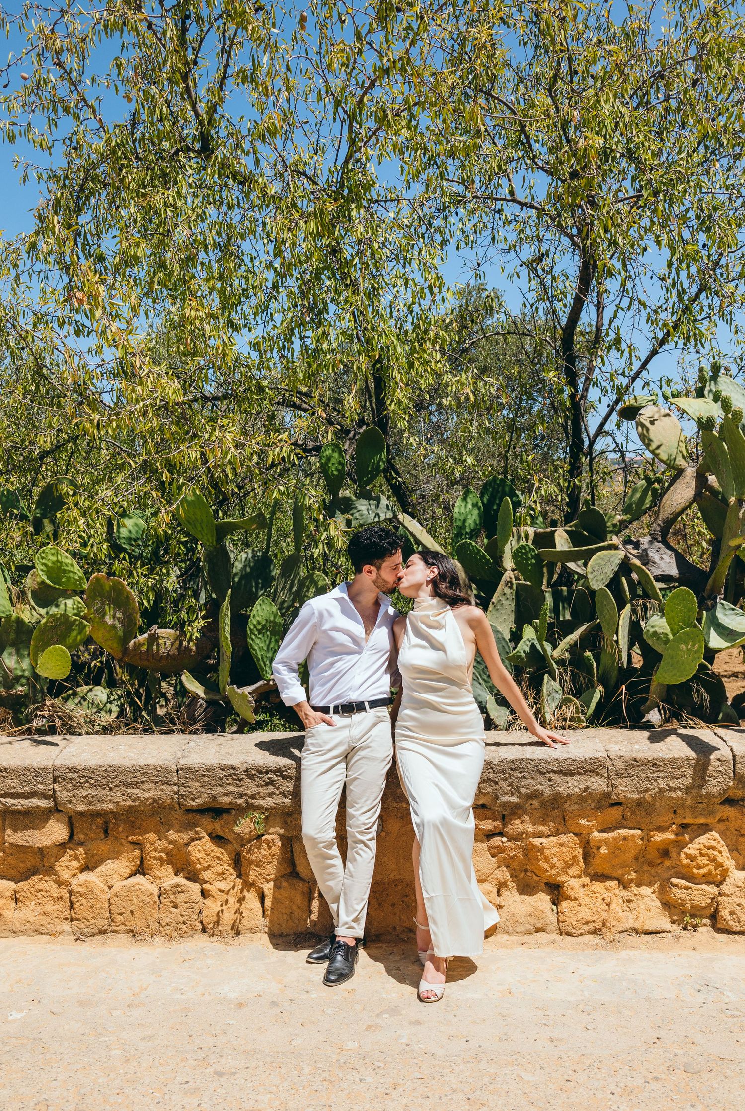 Couple in white outfits share romantic moments against a stone wall and cactus garden backdrop.